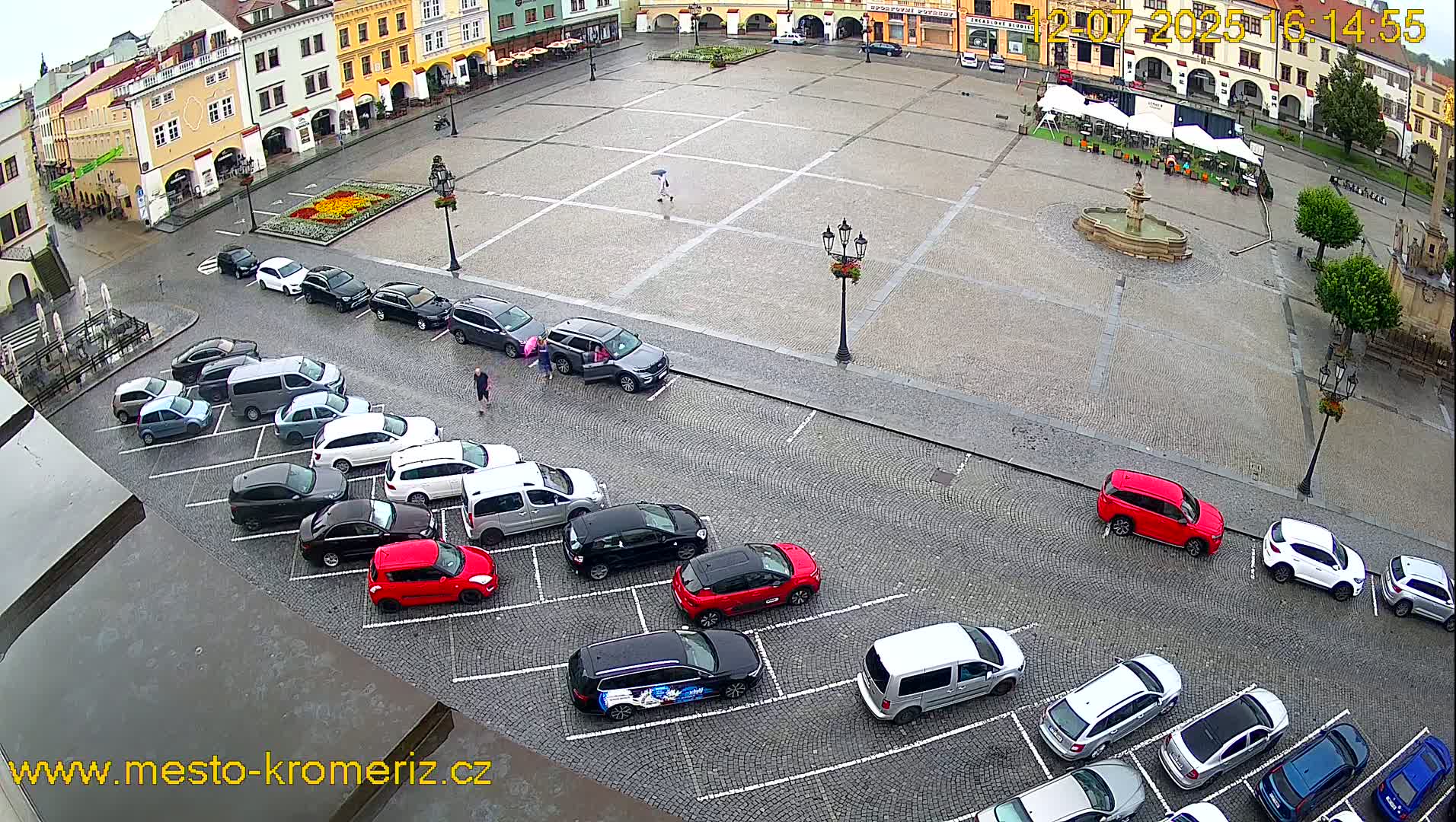 A large town square with a fountain in the center, surrounded by buildings and cars parked along the edges on a sunny day.