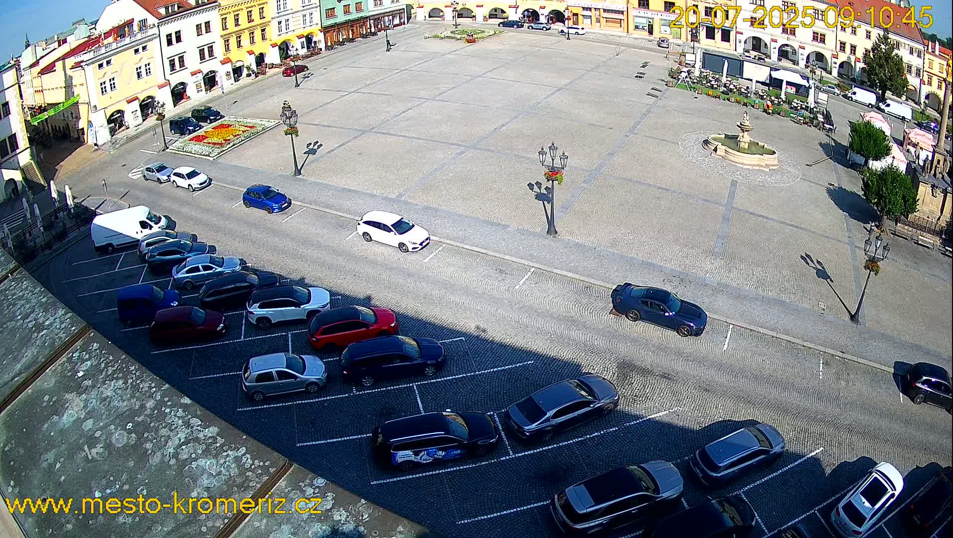 A large town square with a fountain in the center, surrounded by buildings, and several cars parked along the sides on a sunny day.