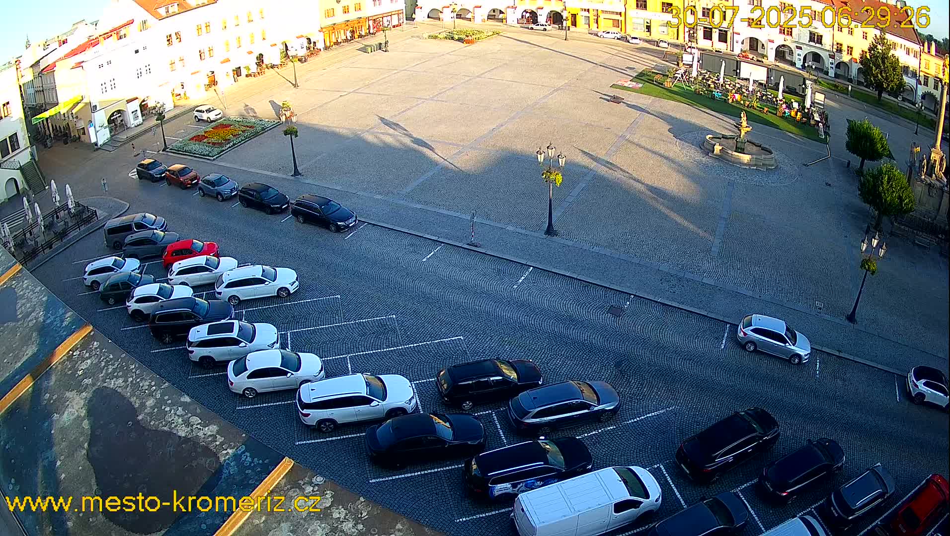 A large town square with a fountain in the center, surrounded by buildings, and several cars parked along the sides on a sunny day.