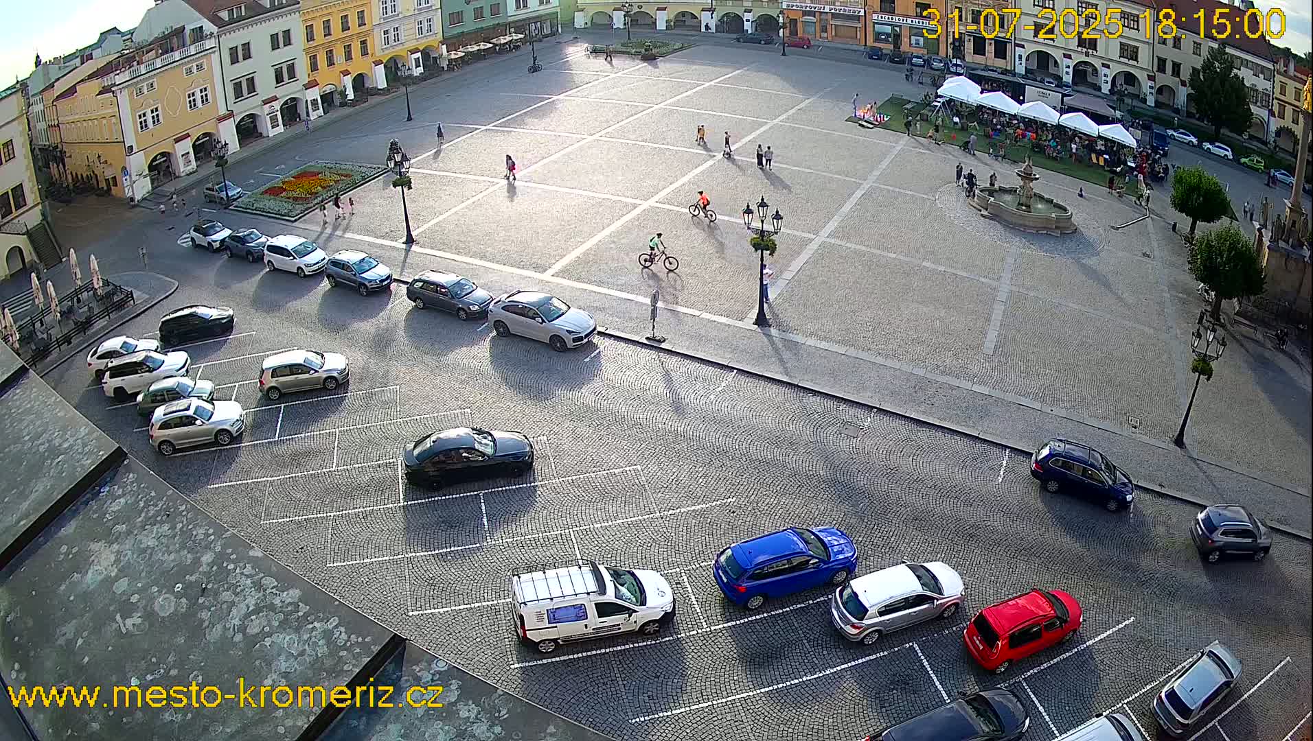 A cobblestone town square with parked cars and a few people walking and biking, under sunny skies.