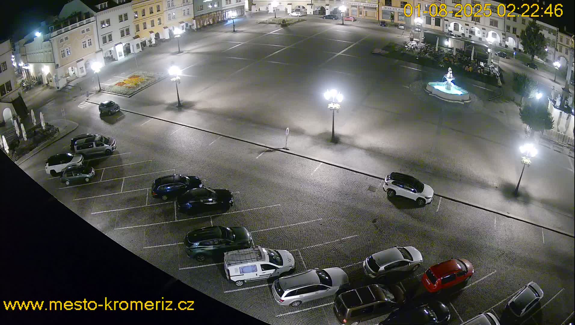 A nighttime aerial view shows a town square with a fountain and several cars parked in a lot surrounding it.