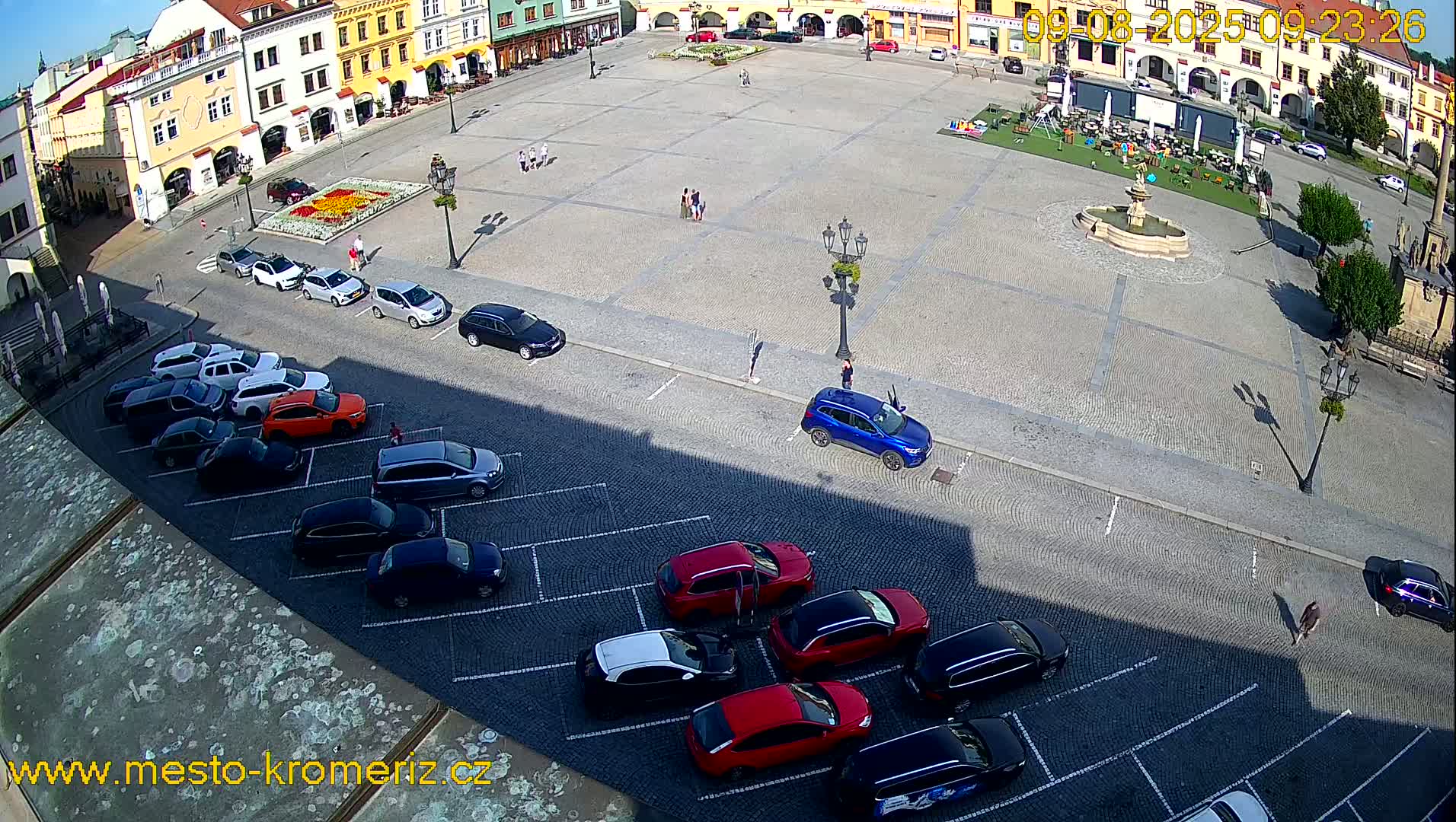 A sunny day reveals a town square with a fountain, parked cars, and a few people walking around, all surrounded by colorful buildings.
