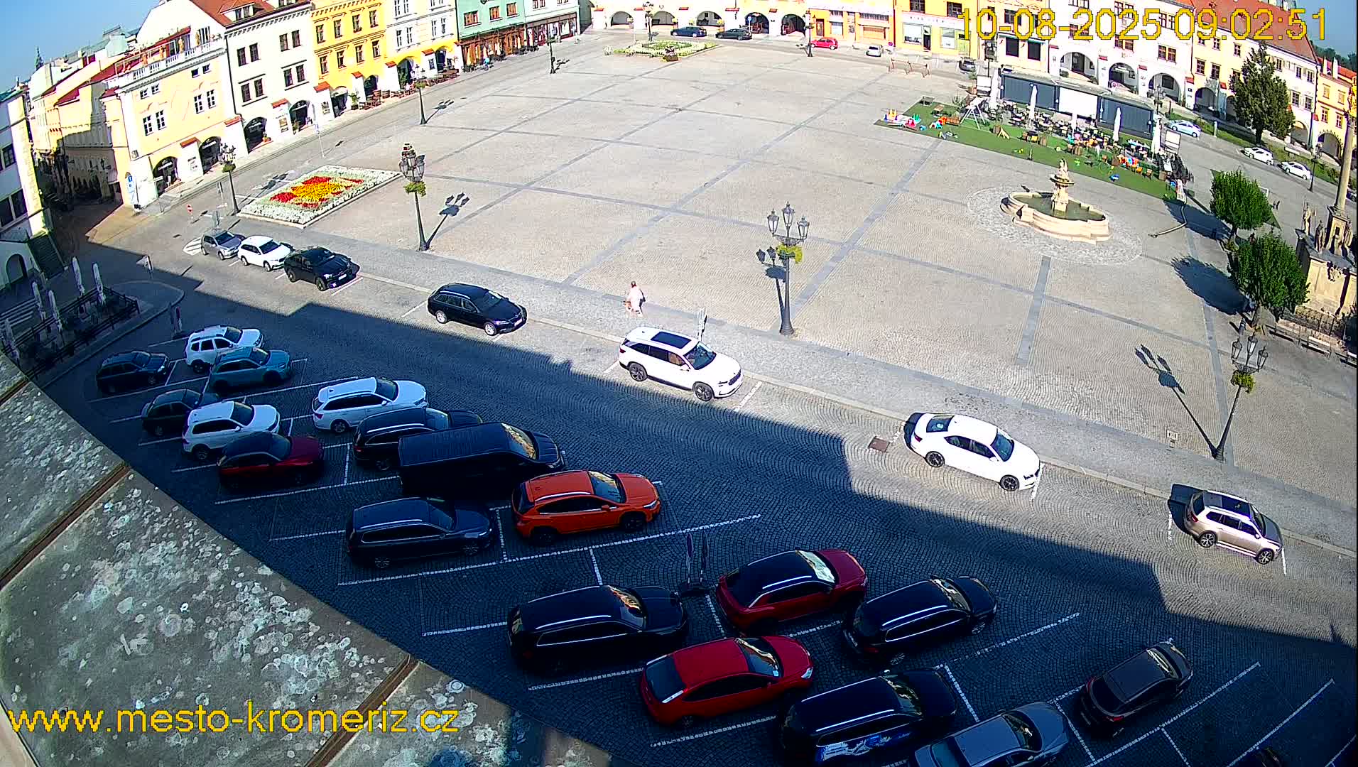 A large town square with a fountain in the center is surrounded by parked cars and buildings on a sunny day.