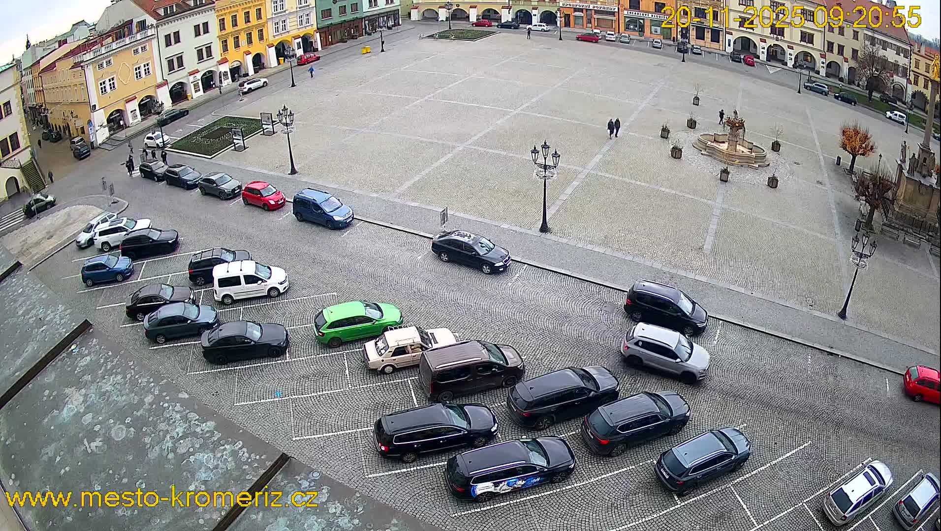 An overcast sky hangs over a European town square featuring colorful historic buildings, numerous parked cars along the paved edges, a central fountain, and a few pedestrians.