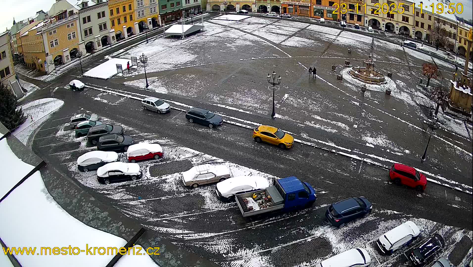A wintry aerial view shows a historic town square and surrounding cobblestone streets partially covered in light snow, with several cars and a few pedestrians visible under an overcast sky.