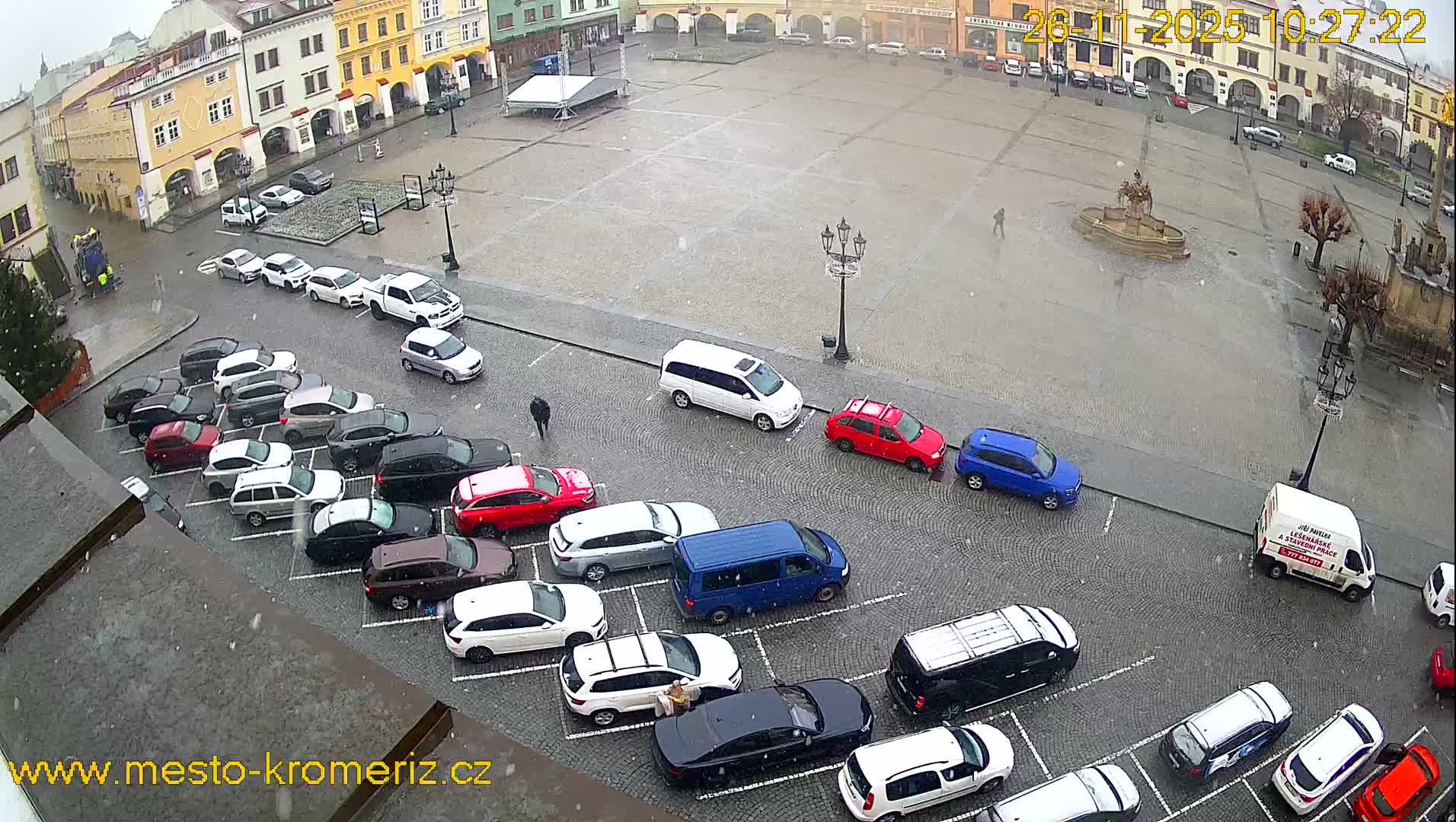 From an elevated view, a wide, wet cobblestone town square is visible under an overcast sky with light snow falling, featuring numerous parked cars, historical buildings lining the perimeter, and a few scattered pedestrians.