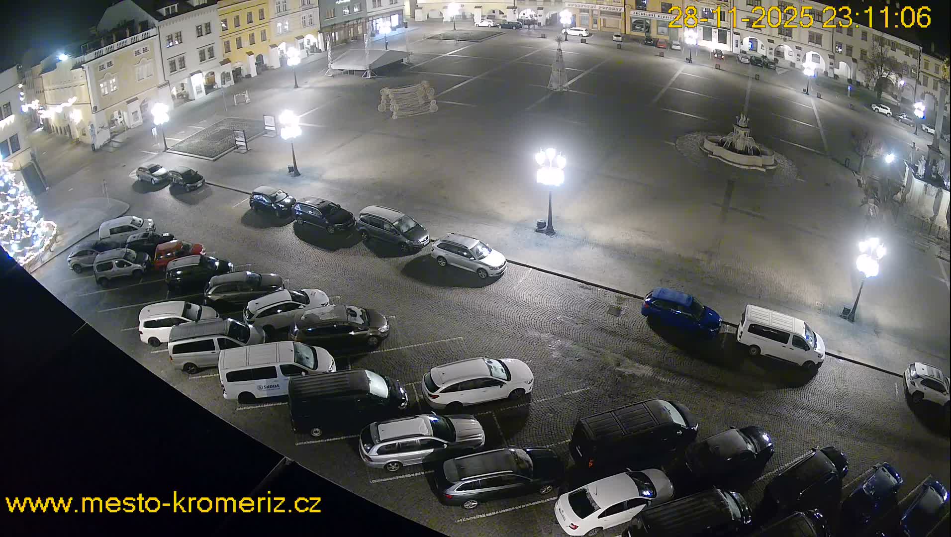 On a clear night, a high-angle view reveals a well-lit European town square filled with parked cars, surrounded by historic buildings and featuring a central fountain and a glowing Christmas tree.
