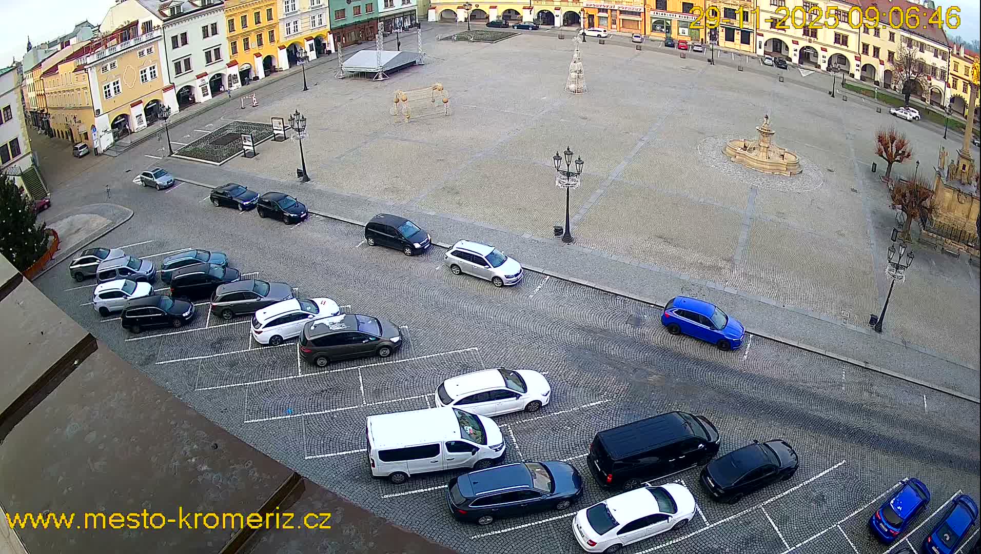 An overhead view reveals a European town square adorned with historic buildings, a central fountain, festive structures, and numerous parked cars on an overcast day.
