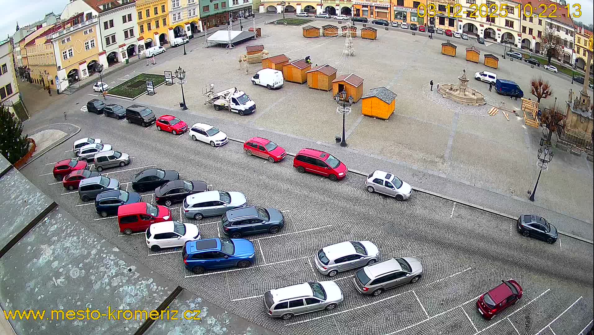 An overhead view reveals a bustling European town square lined with historic buildings, featuring numerous parked cars, several market stalls being set up around a central fountain, all under an overcast sky.