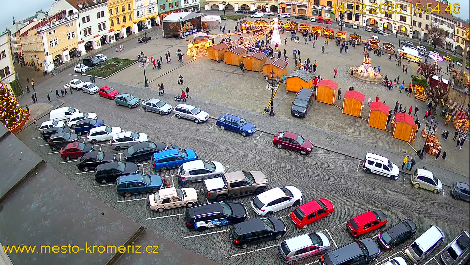 An overhead view reveals a bustling European town square lined with historic buildings, featuring numerous parked cars, several market stalls being set up around a central fountain, all under an overcast sky.