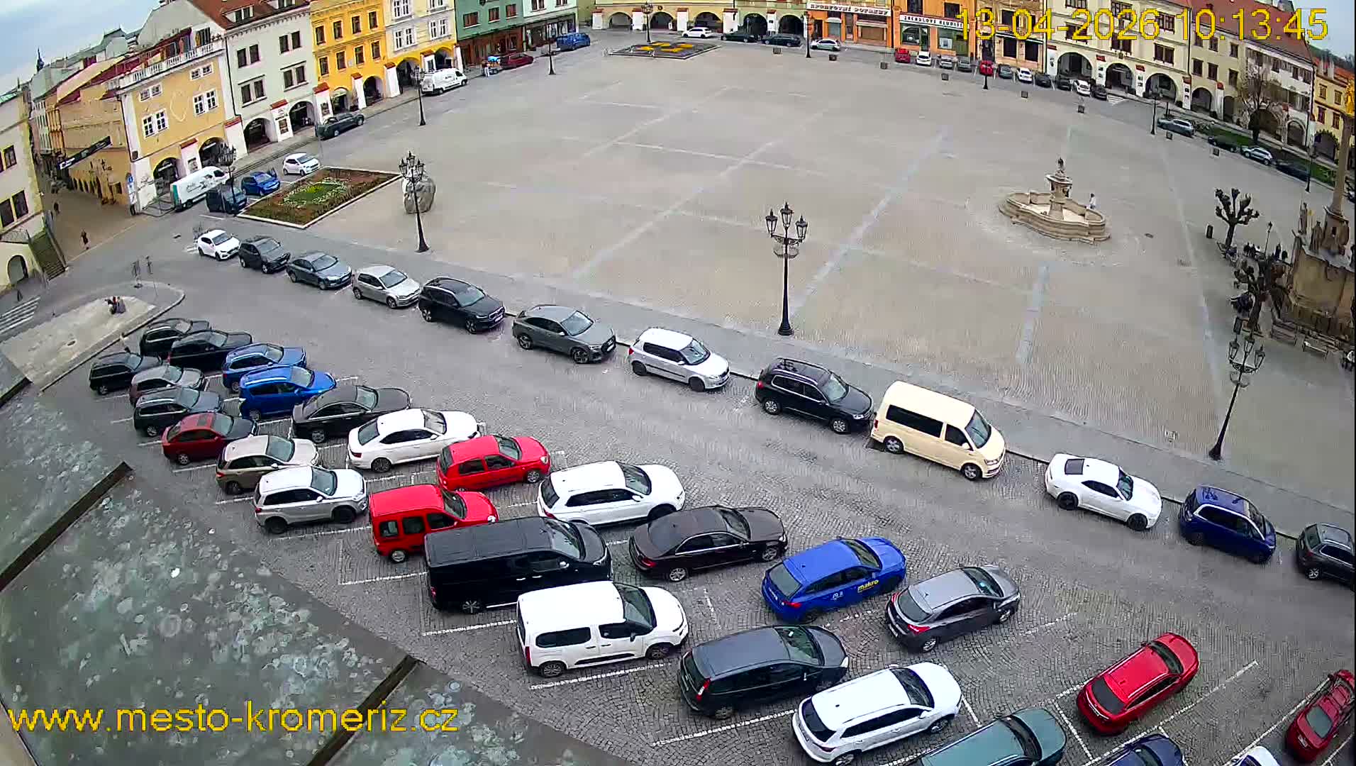 An overhead view reveals a bustling European town square lined with historic buildings, featuring numerous parked cars, several market stalls being set up around a central fountain, all under an overcast sky.