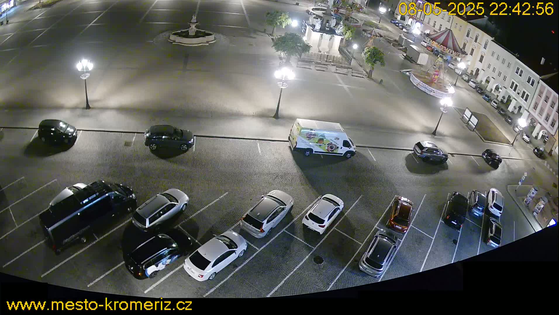 A nighttime aerial view of a town square's parking lot, filled with cars, and a van, under artificial light.