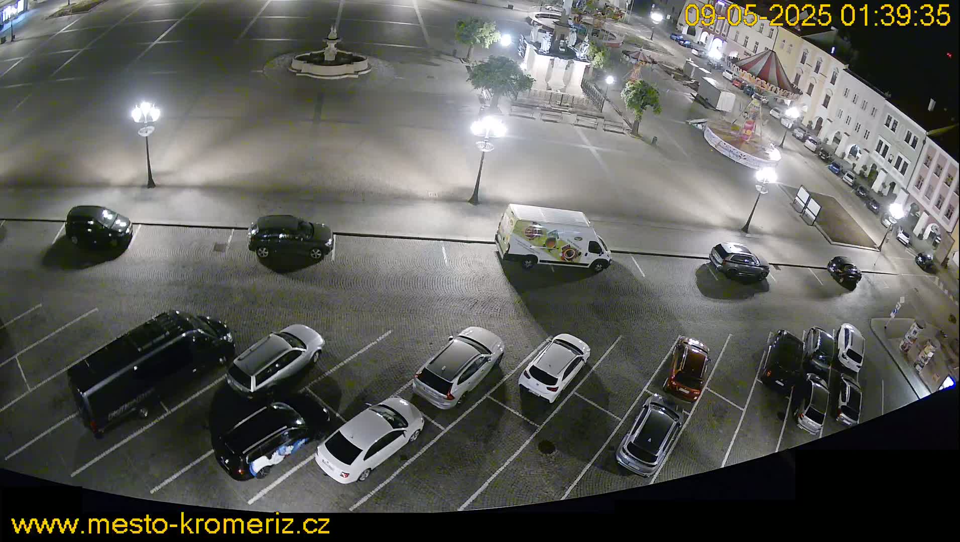 A nighttime, high-angle view of a town square with cars parked in a lot surrounding it, and a fountain in the center of the square.
