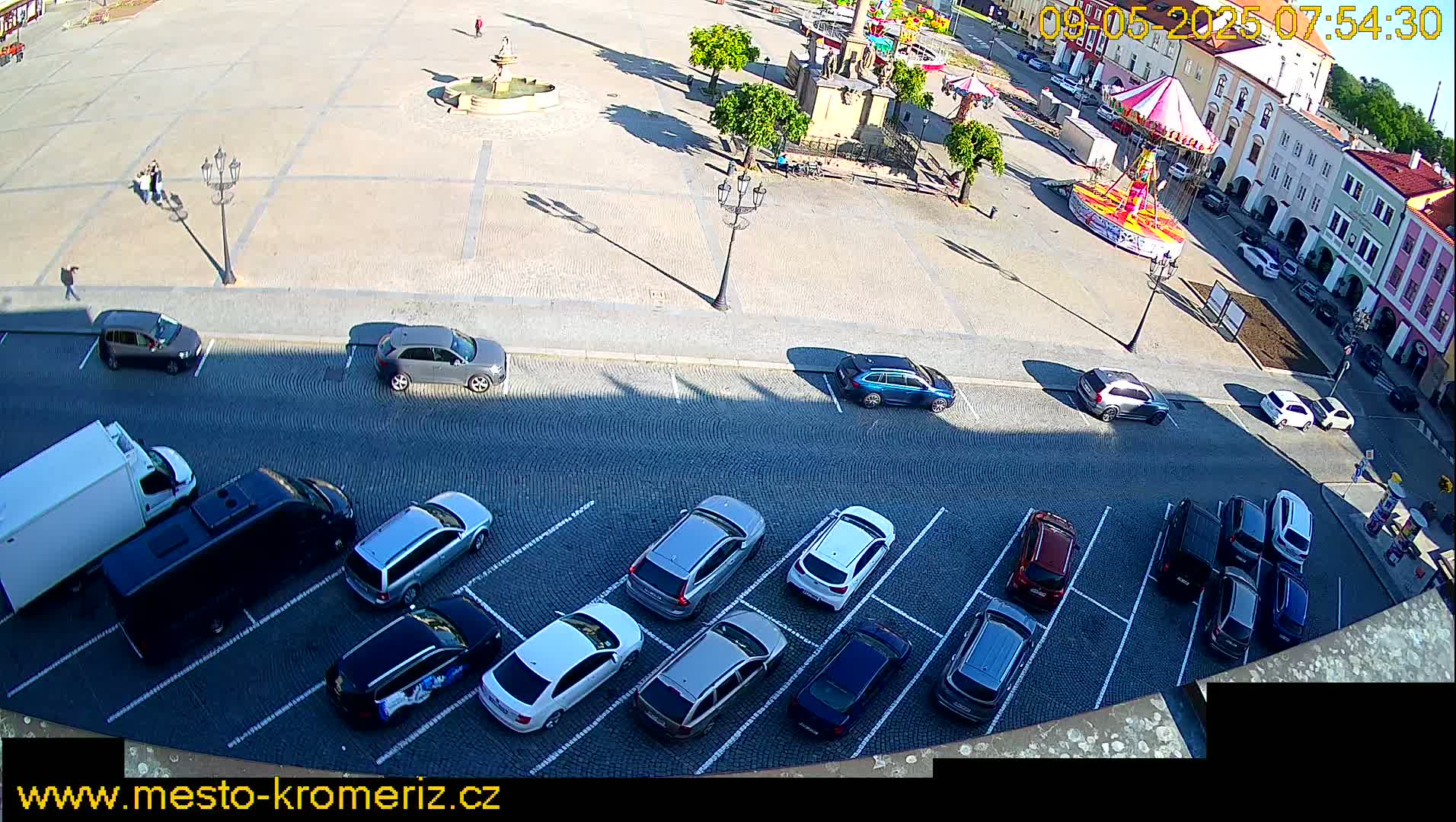 A sunny day shows a town square with a carousel, a fountain, and many cars parked along the street.