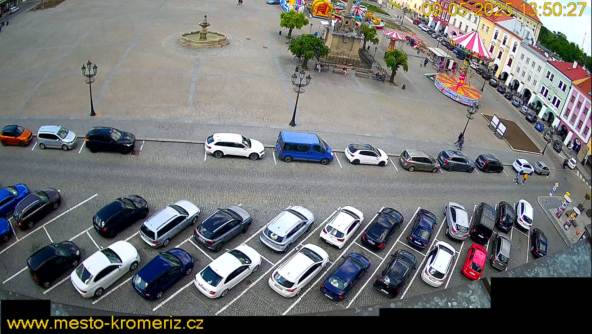 A paved town square with a fountain and carousel, surrounded by parked cars under a partly sunny sky.