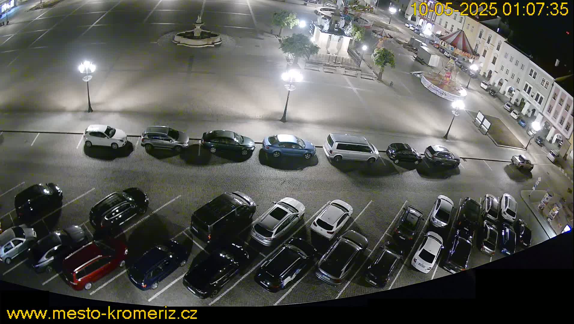 A nighttime overhead view of a town square parking lot filled with numerous cars under artificial lights.