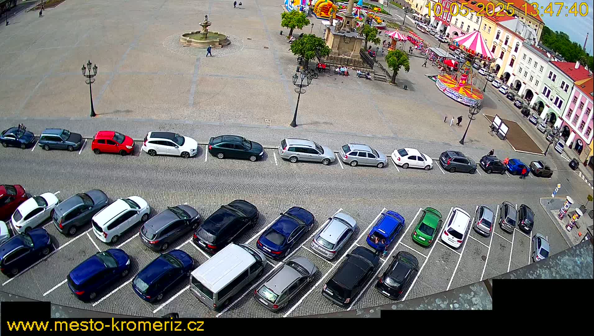 A town square with a carousel, fountain, and numerous cars parked in two rows, on a sunny day.