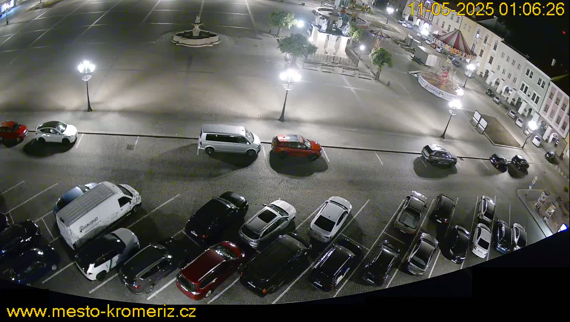 A nighttime, overhead view of a town square with several cars parked in a lot, and a fountain in the center.