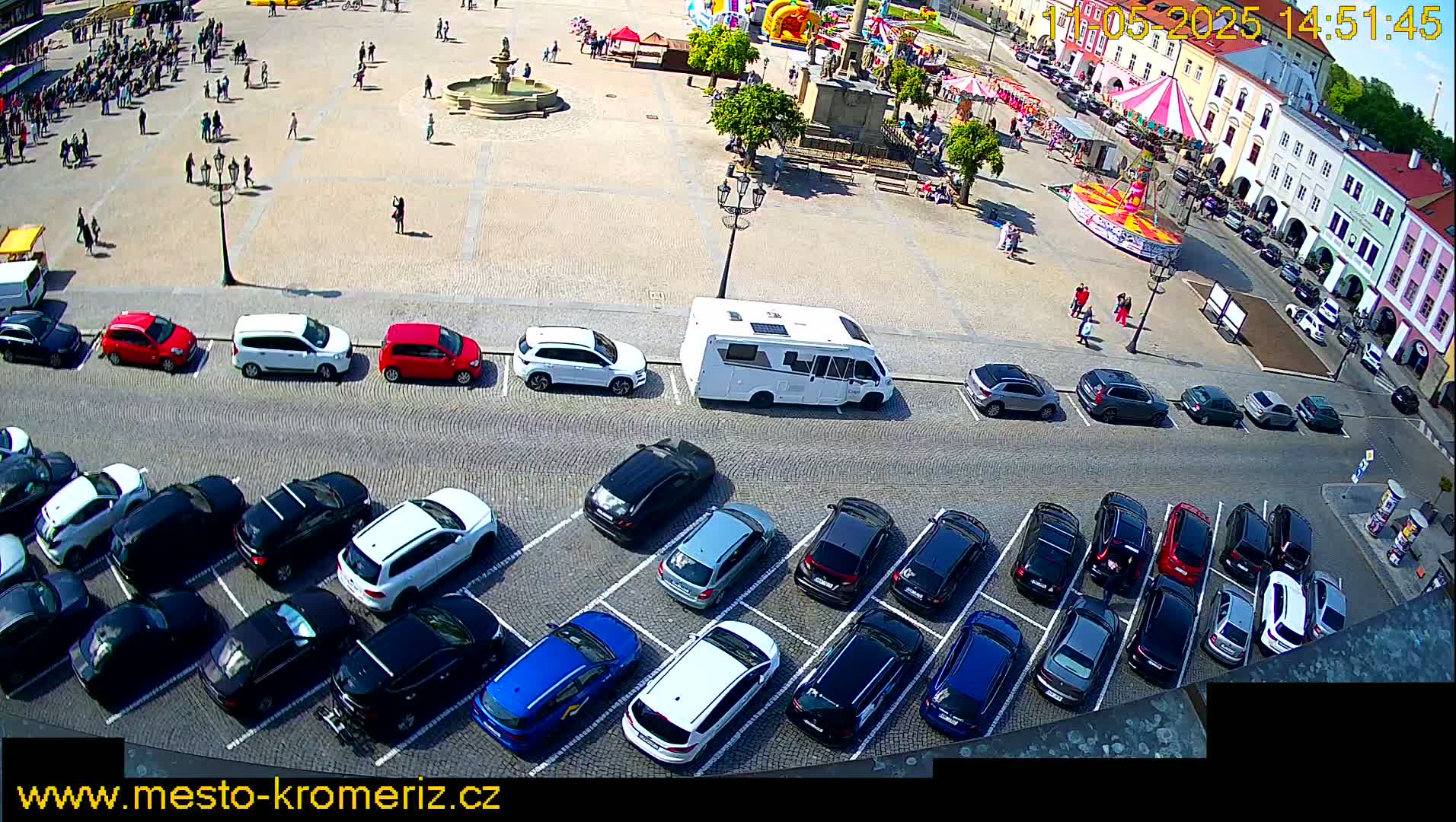 A sunny town square with a fountain, a carousel, and numerous cars parked in a lot.