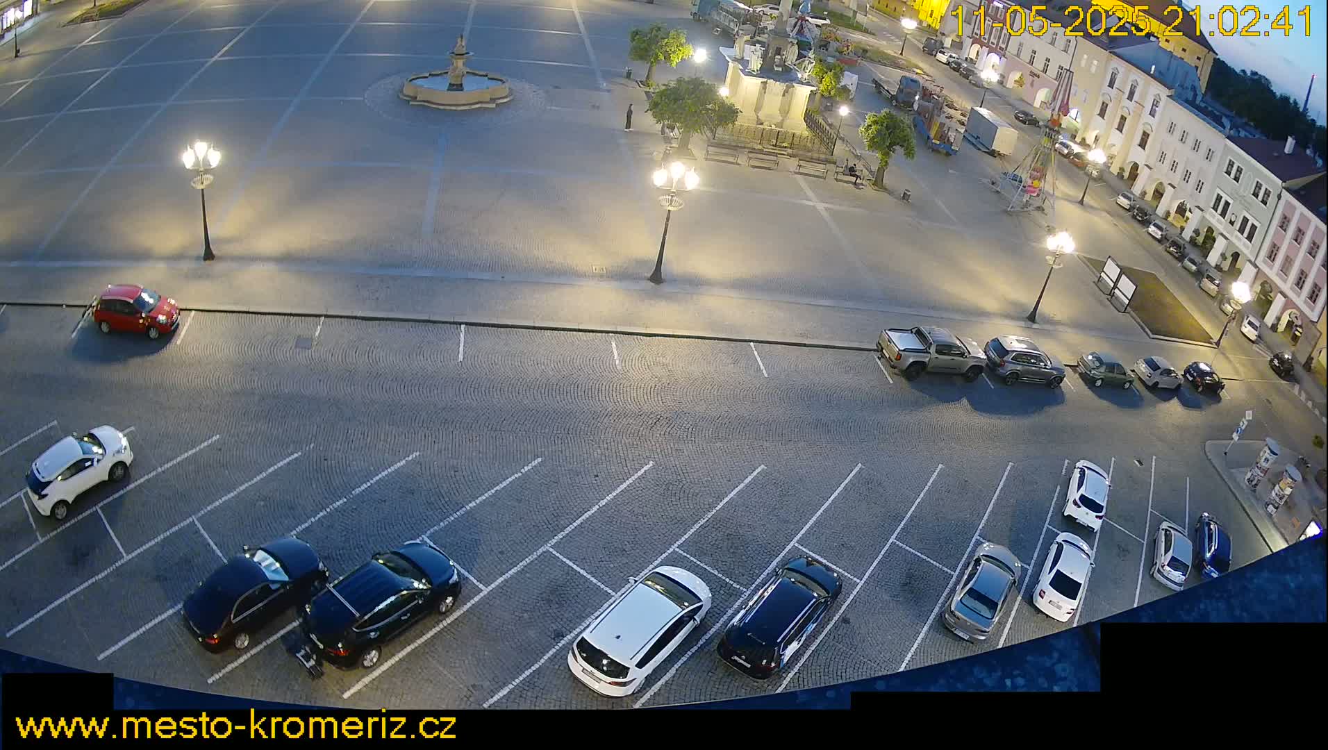 A nighttime view of a town square with several cars parked in a lot surrounding it, illuminated by streetlights.