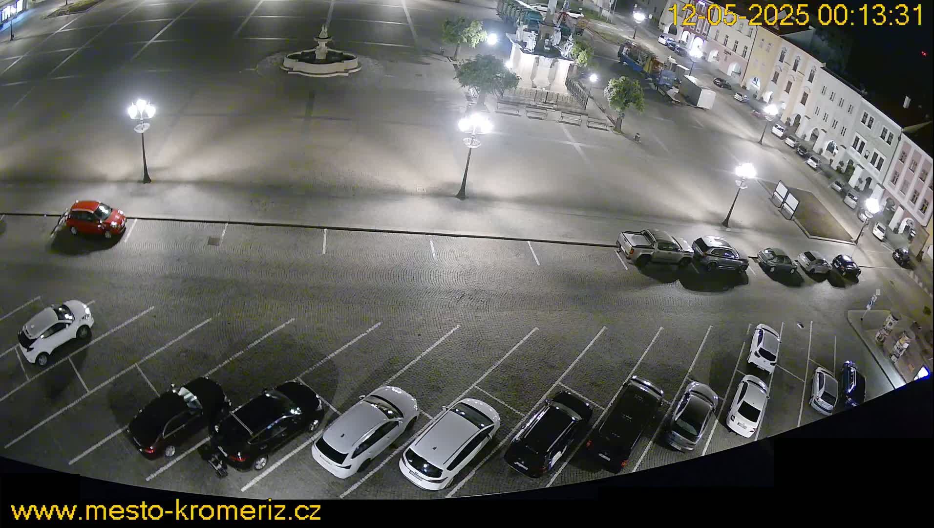 A nighttime, high-angle view of a town square's parking lot with several cars parked, and a fountain and statue visible in the center of the square.