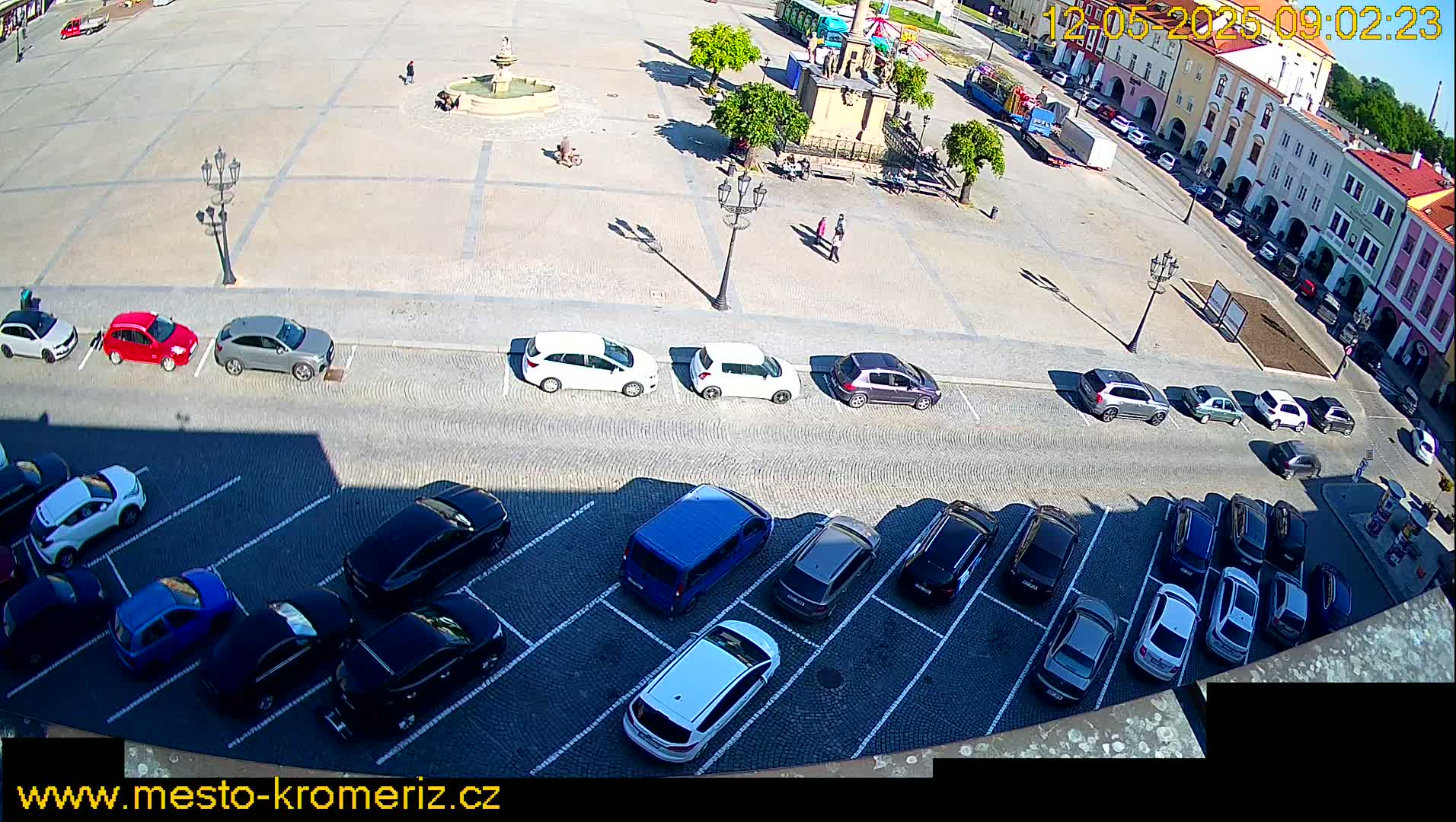 A sunny day reveals a town square with a fountain and numerous cars parked in a lot and along the street, next to buildings.