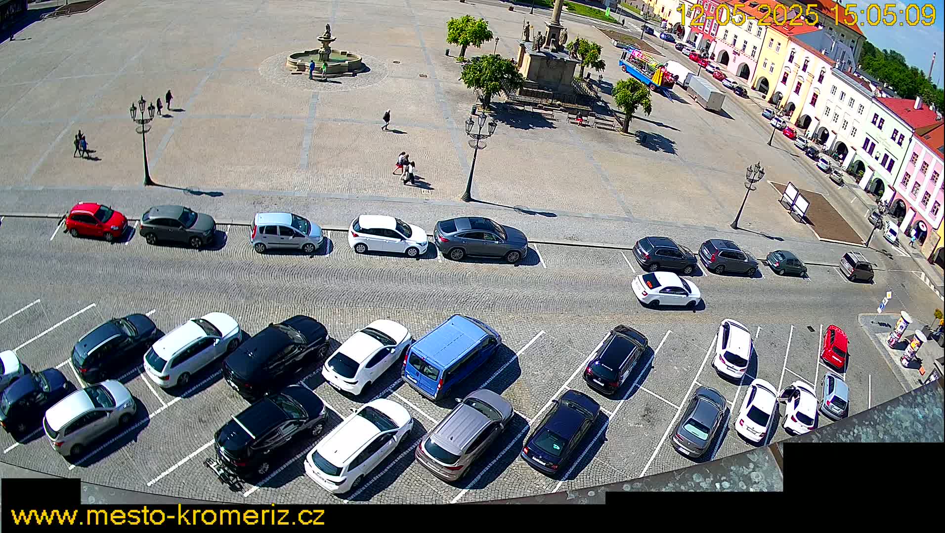 A town square with a fountain and several parked cars under a sunny sky.