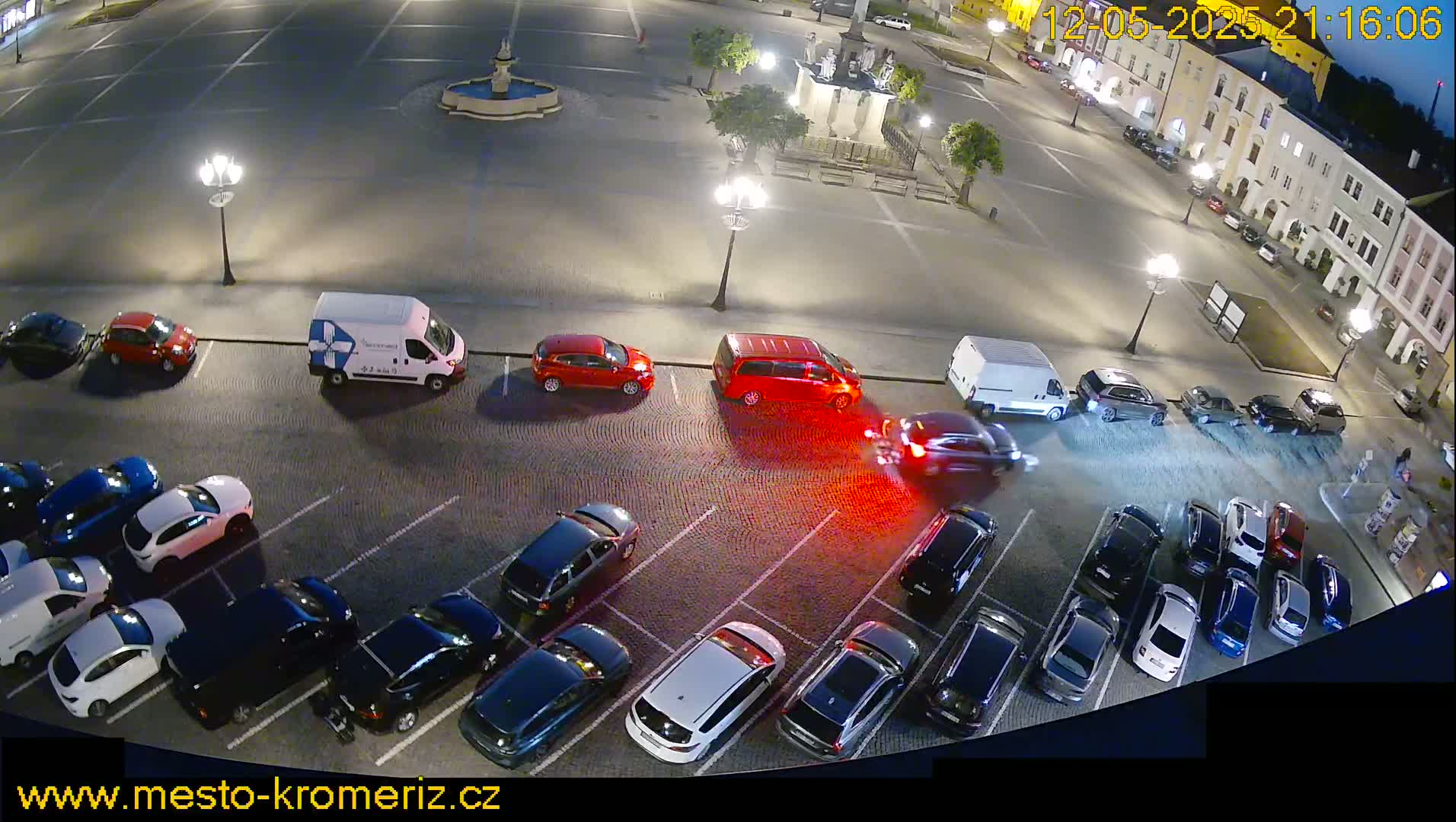 At night, numerous cars are parked in a large square, with a few vehicles driving through.