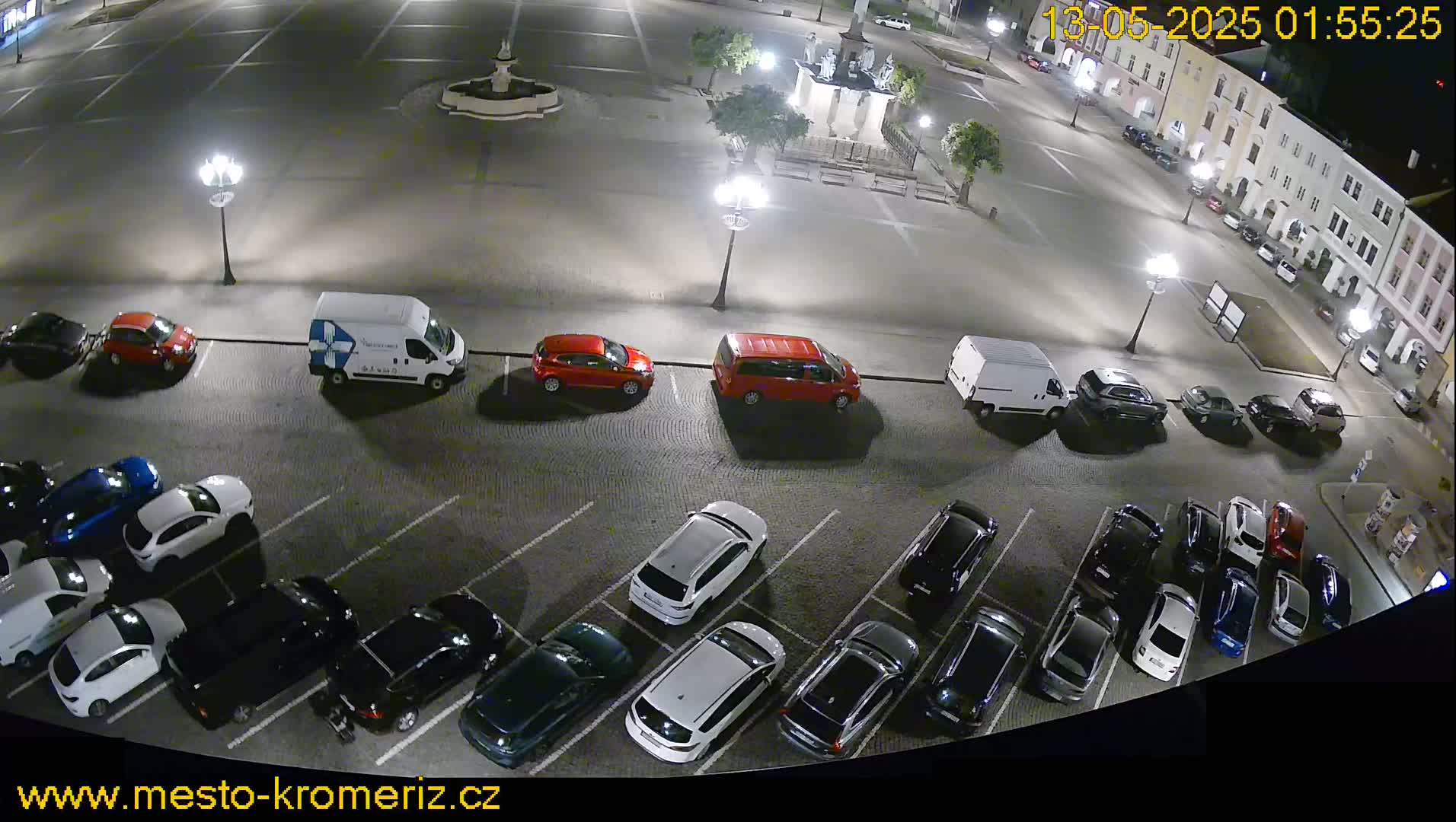 A nighttime aerial view of a mostly empty parking lot next to a town square, with several cars parked and streetlights illuminating the scene.