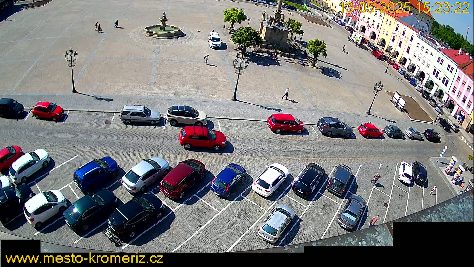A sunny day shows a town square with a fountain and several cars parked in a lot surrounding the square, with some pedestrians walking.