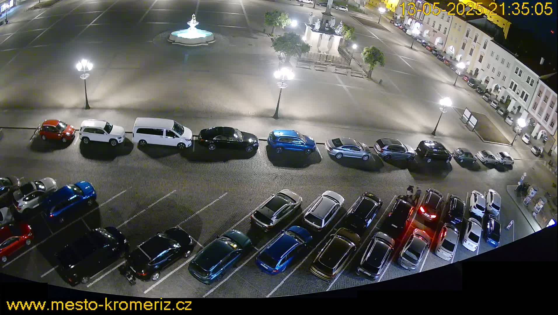 A nighttime view of a town square with many cars parked in a lot, and a fountain in the center.