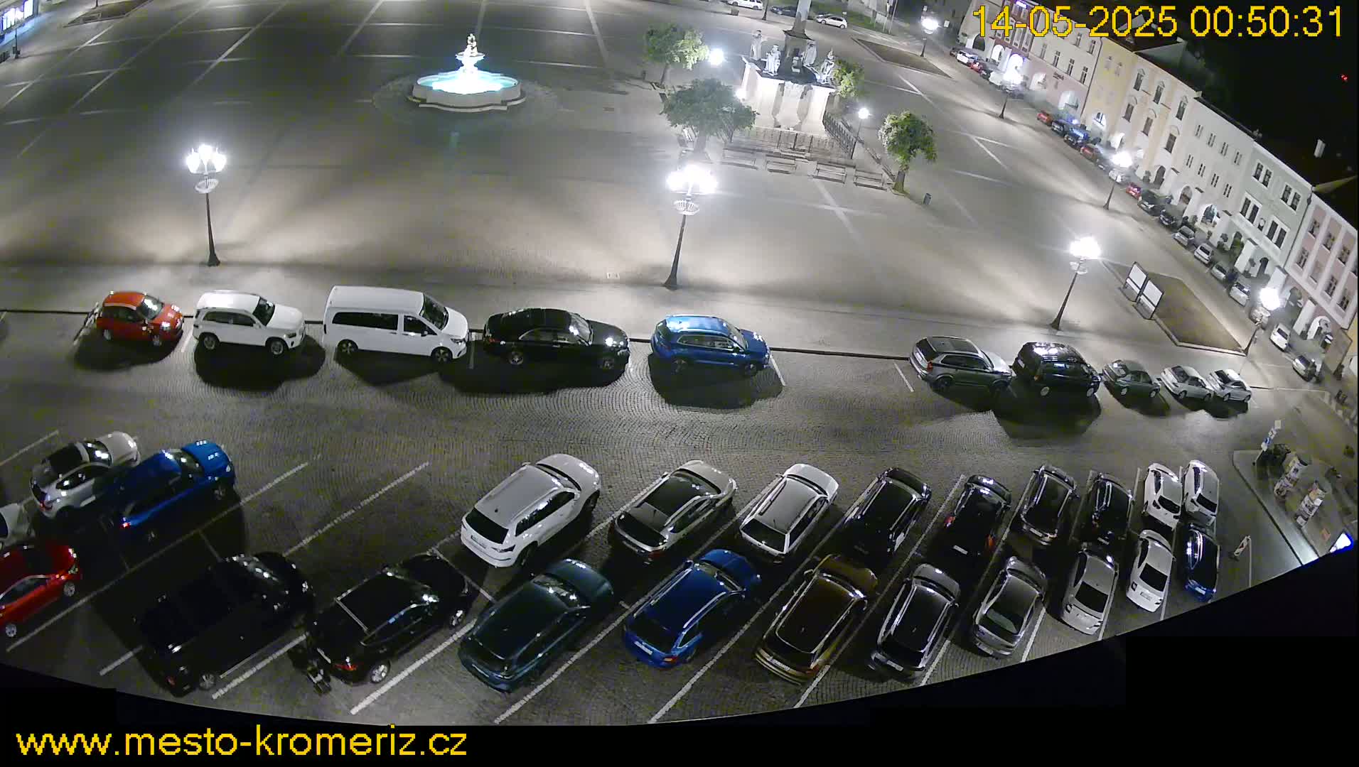 A nighttime aerial view of a town square shows numerous cars parked in a lot, with a fountain and a building visible in the background.