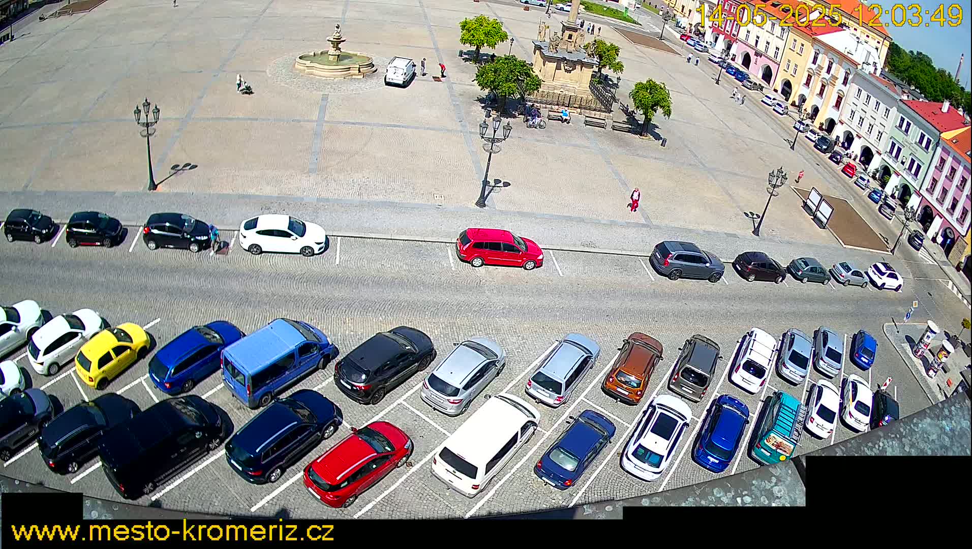 A town square with a fountain and parked cars is viewed from above on a sunny day.
