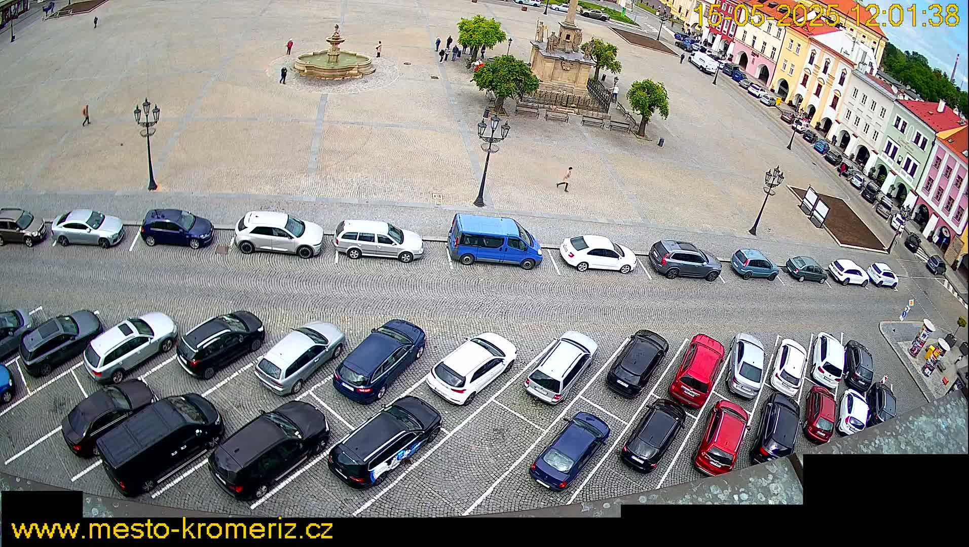 Rows of parked cars fill a cobblestone town square surrounding a fountain and a stone monument, under a partly cloudy sky.