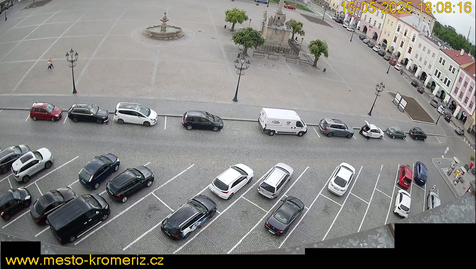 A paved town square with a fountain and parked cars, viewed from above on an overcast day.
