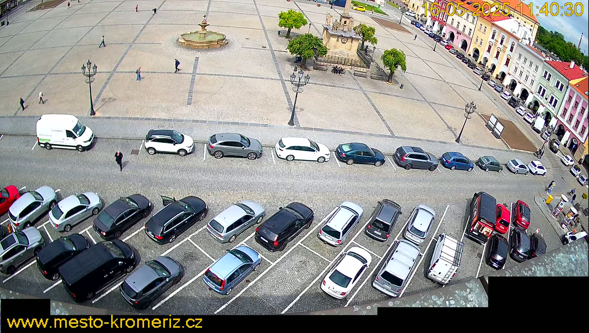 A high-angle view shows a town square with a fountain, several parked cars, and a few pedestrians on a sunny day.