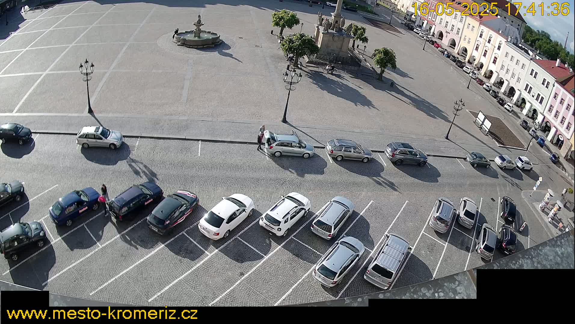 A sunny day shows a town square with a fountain and parked cars in a lot adjacent to the square.