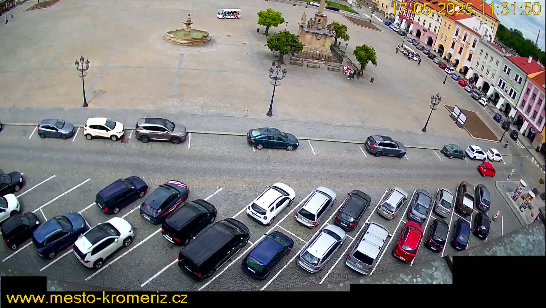 A town square with a fountain and several parked cars, viewed from above on a partly sunny day.