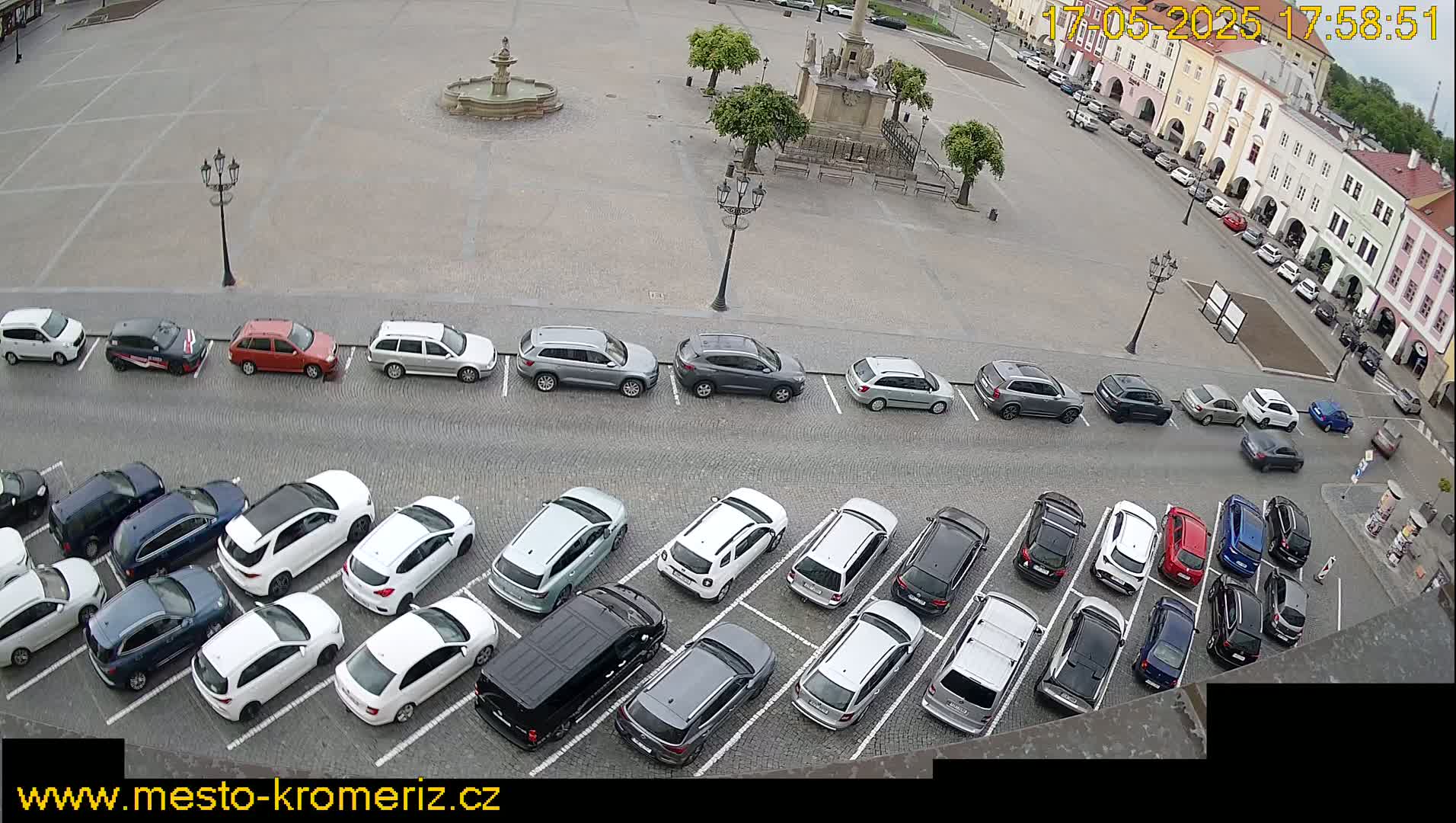 An overcast day reveals a large town square with a fountain and many cars parked in rows, surrounded by buildings.