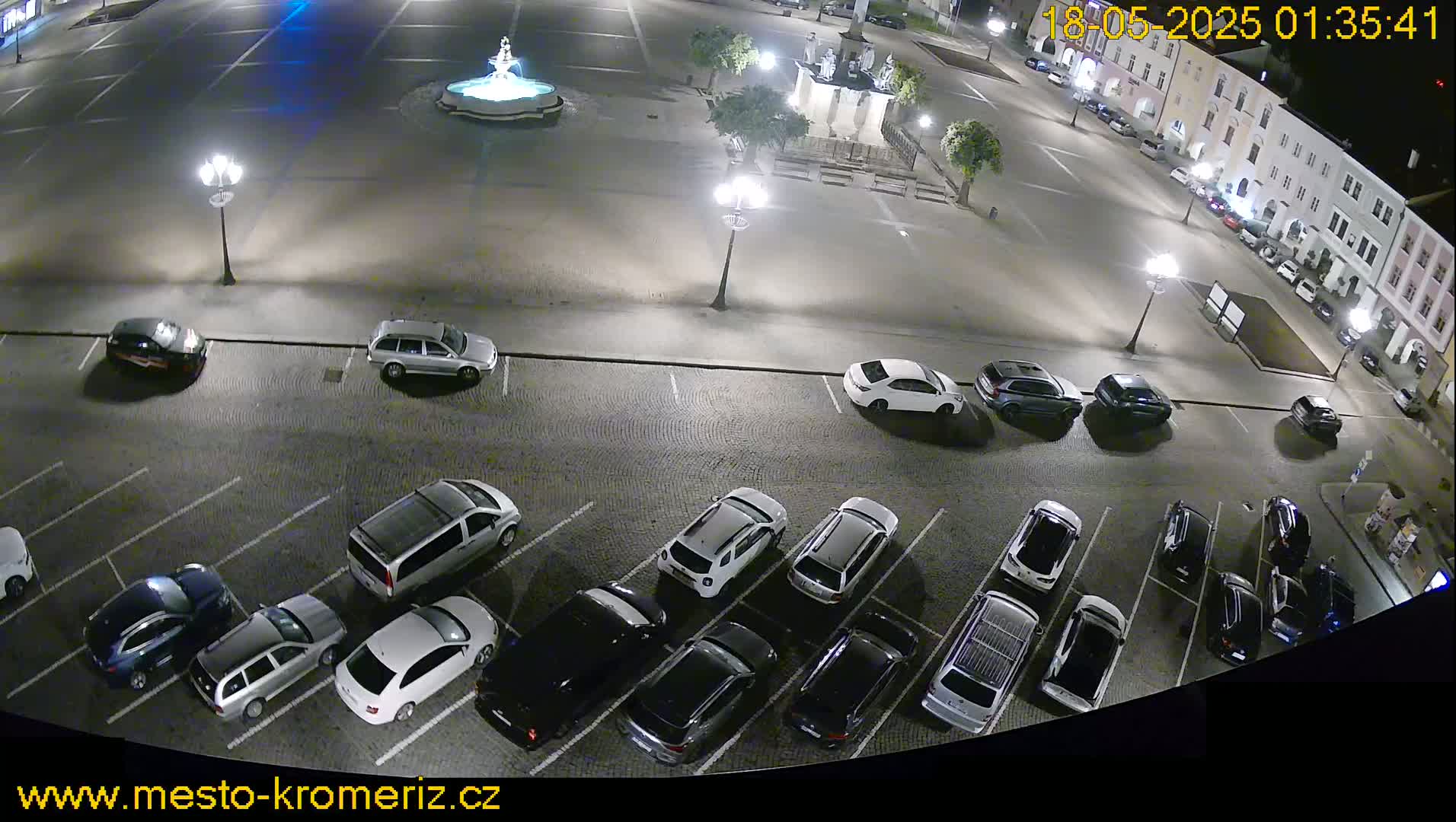 A nighttime, high-angle view of a town square with a fountain and several cars parked in the surrounding lot.