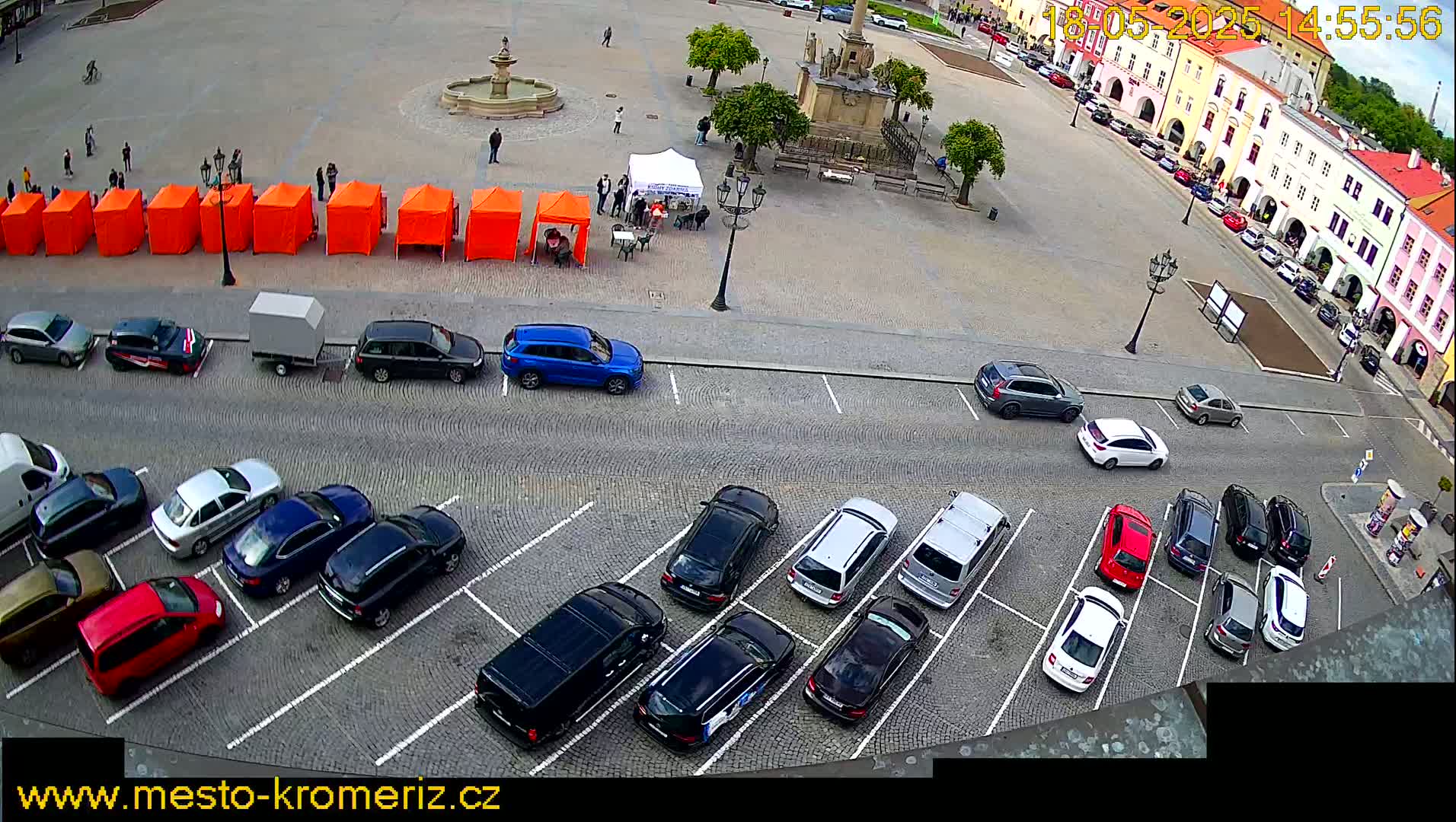 A town square with a fountain, a row of orange tents, and numerous cars parked in a lot, on a partly sunny day.
