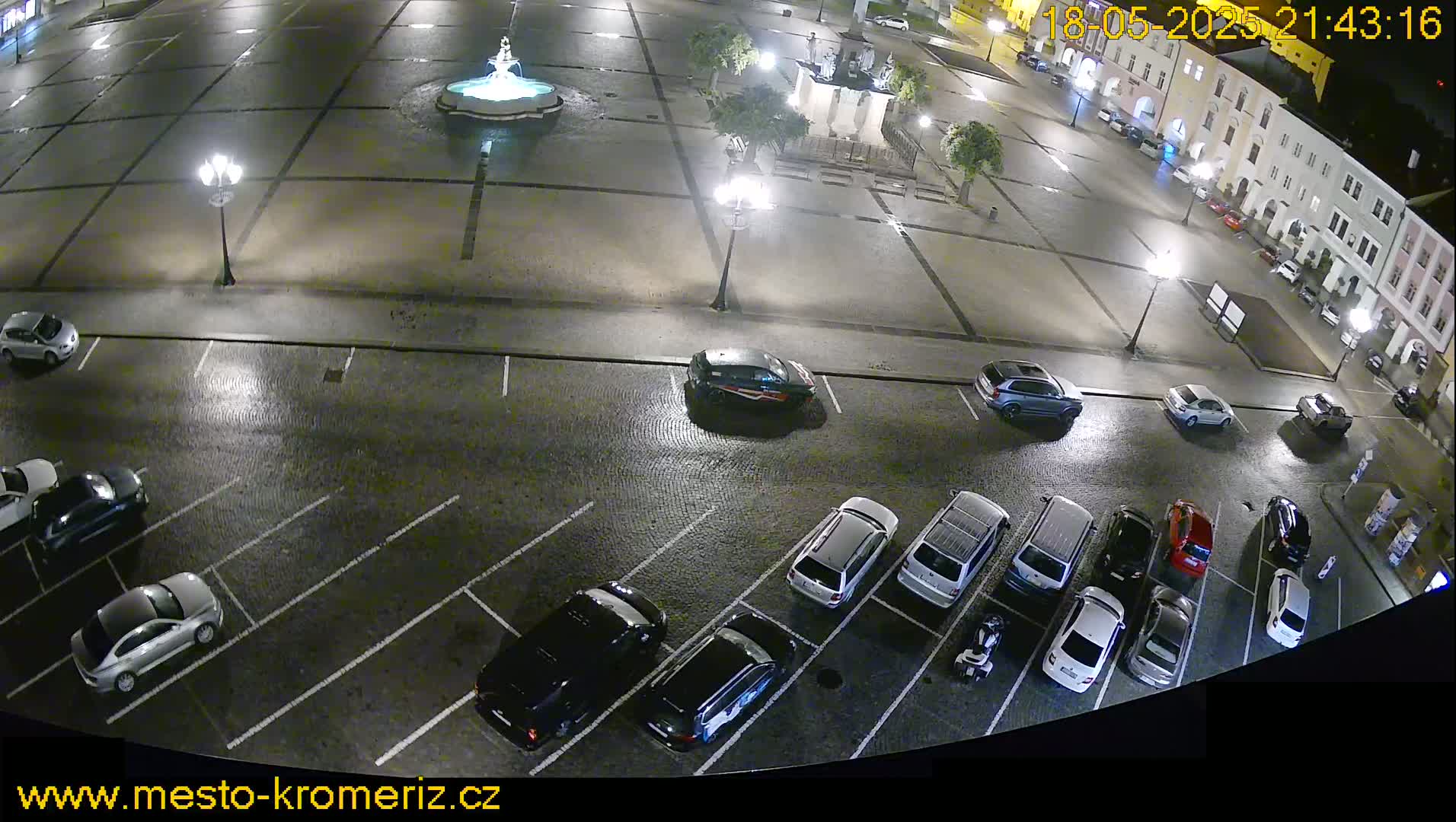 A nighttime, high-angle view of a town square with a fountain, surrounded by buildings and a parking lot filled with cars, under slightly wet conditions.