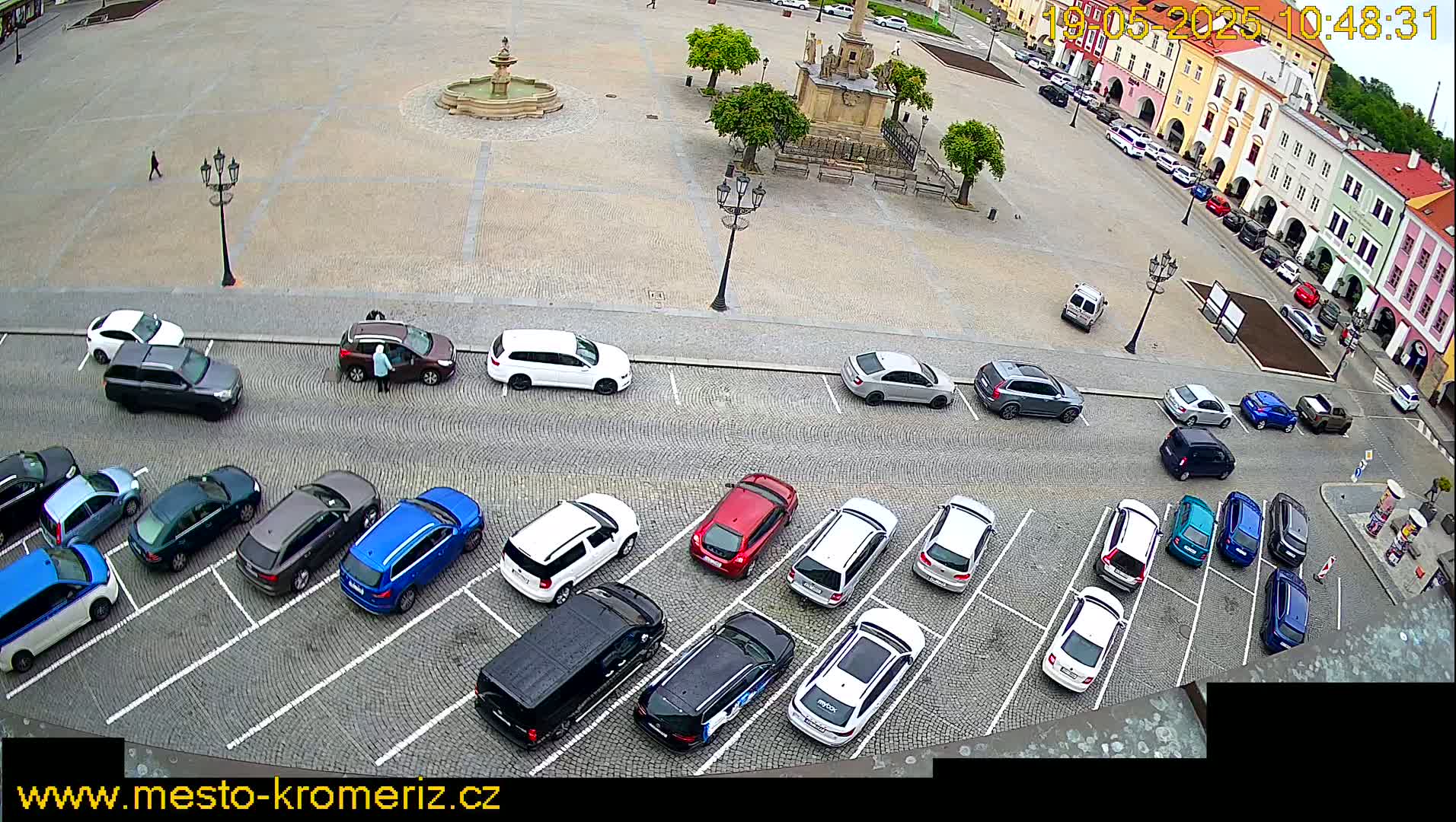 A paved town square with a fountain is surrounded by parked cars and colorful buildings, under an overcast sky.