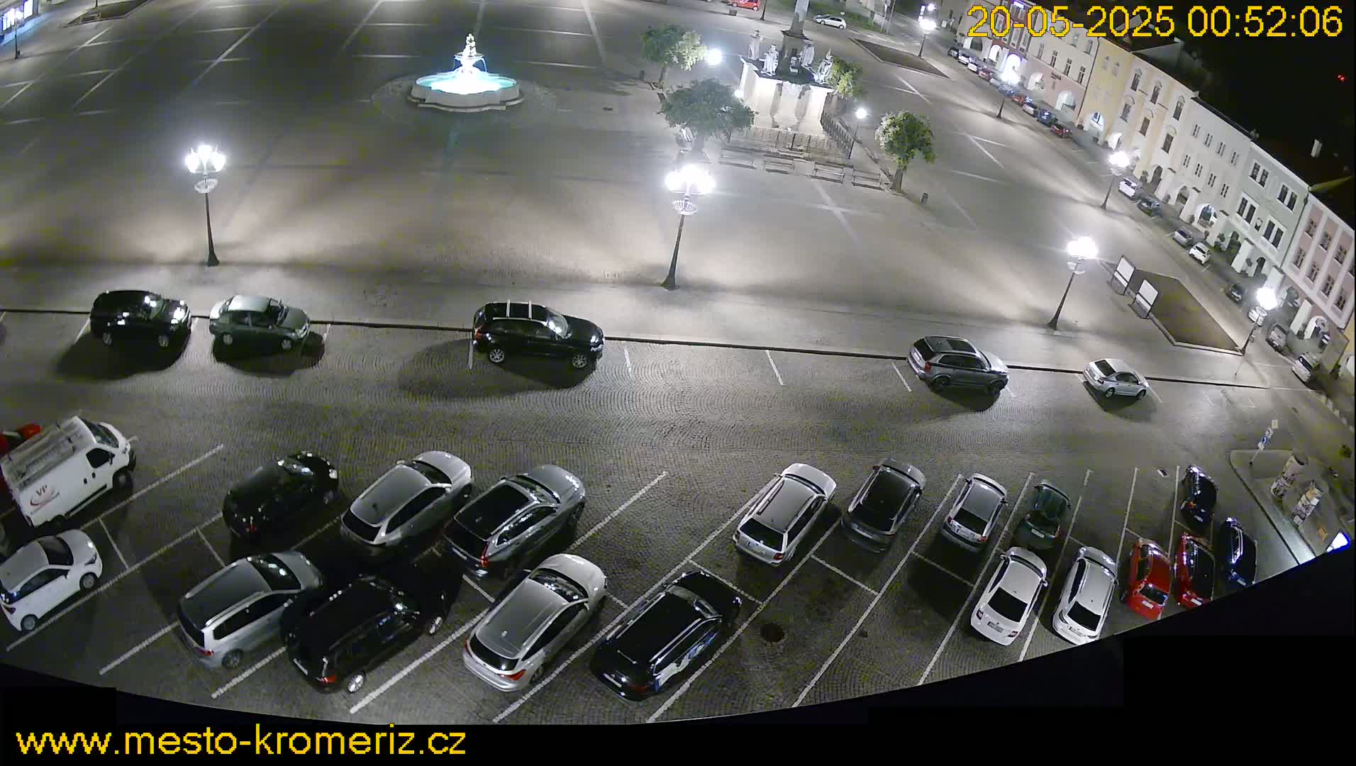 A nighttime aerial view of a town square with a fountain and surrounding buildings, showing numerous cars parked in a lot adjacent to the square.