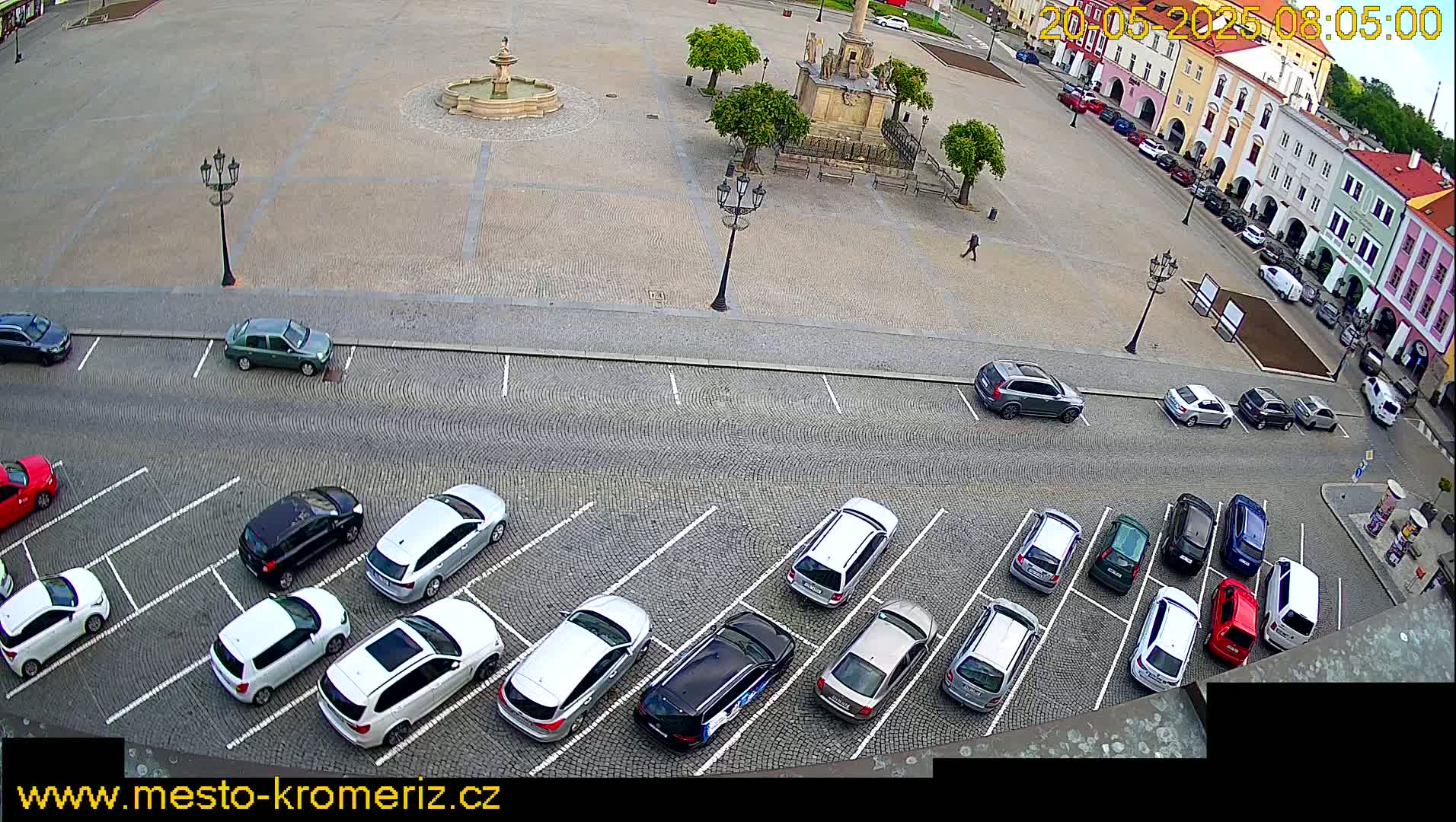 A cobblestone town square with a fountain and several parked cars surrounds it on a mostly sunny day.