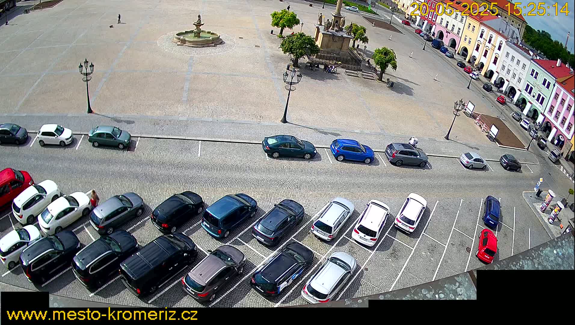 A town square with a fountain and a monument is surrounded by buildings and a parking lot filled with cars on a sunny day.