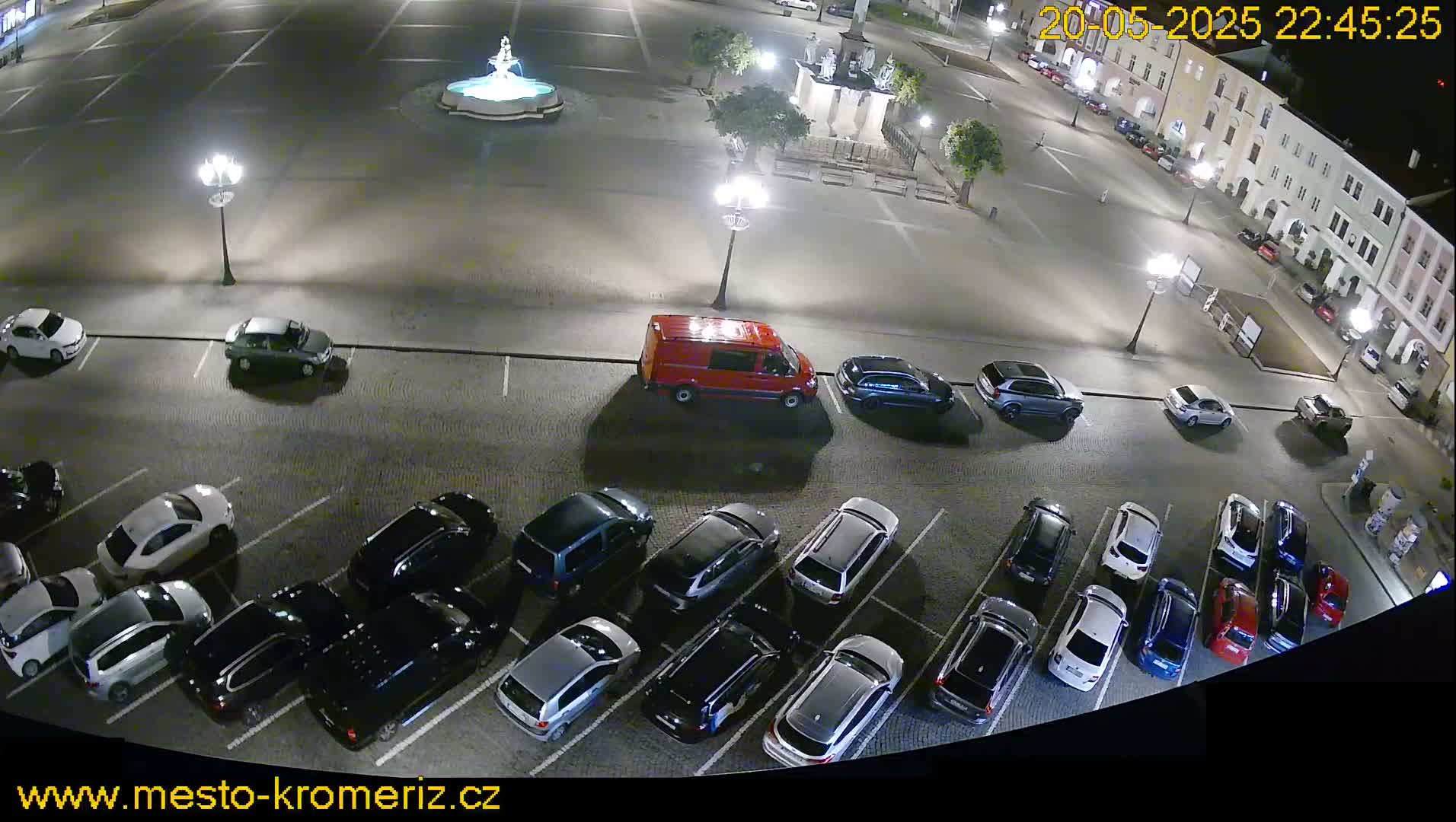 A nighttime, high-angle view of a town square with a fountain and numerous cars parked in a lot surrounding it.