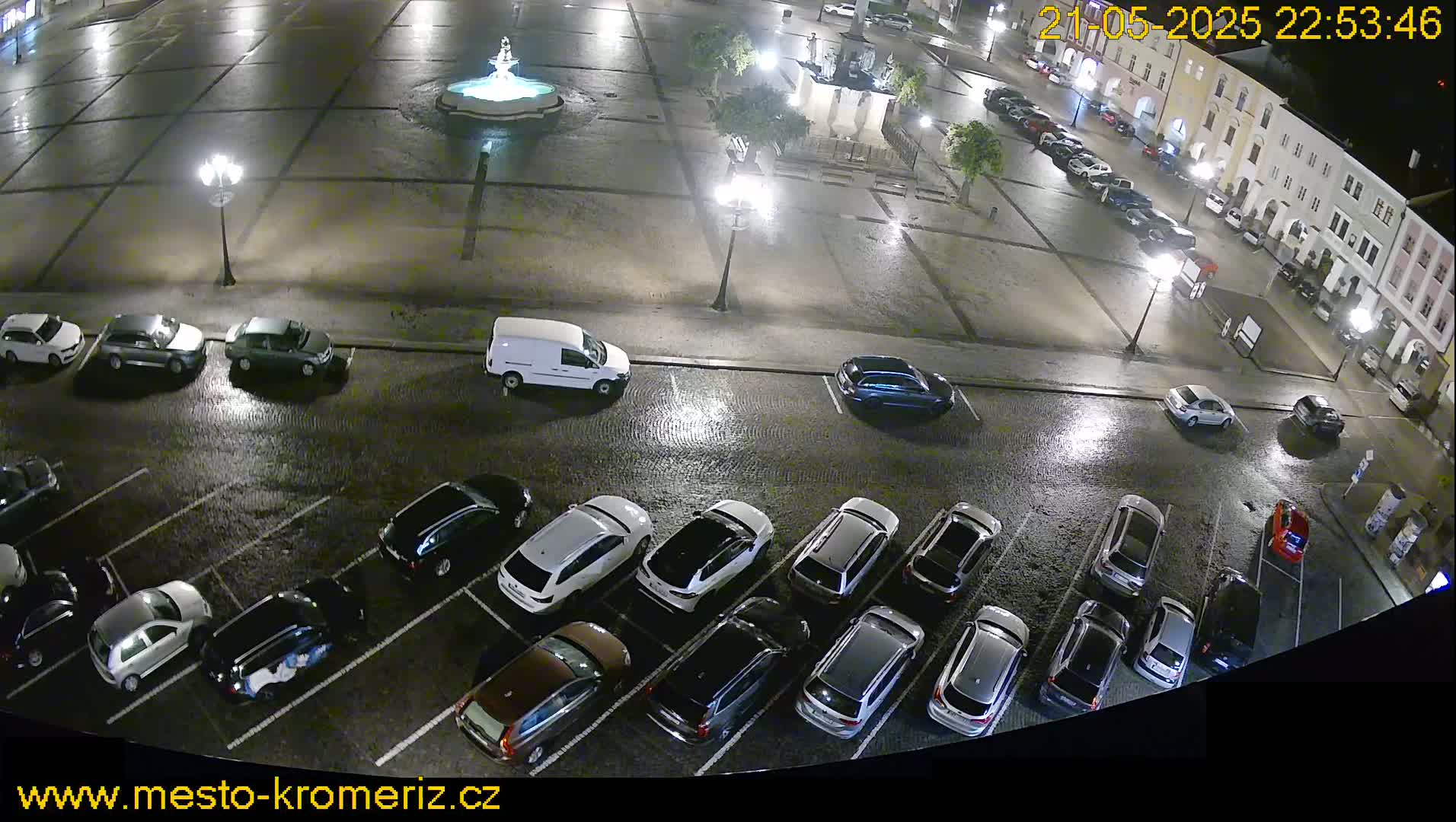 A wet, nighttime view of a town square with a fountain and numerous cars parked in a lot adjacent to it.