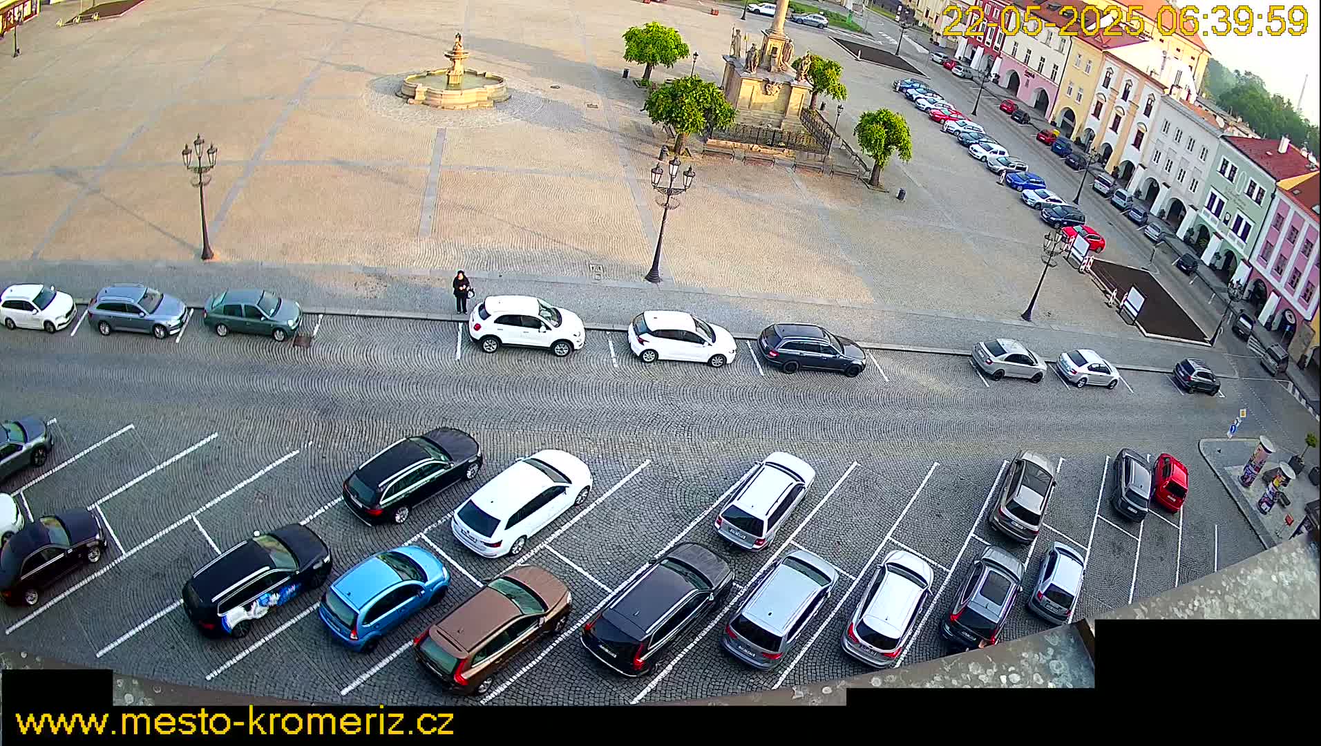 A cobblestone town square with a fountain and several parked cars, under a mostly sunny sky.