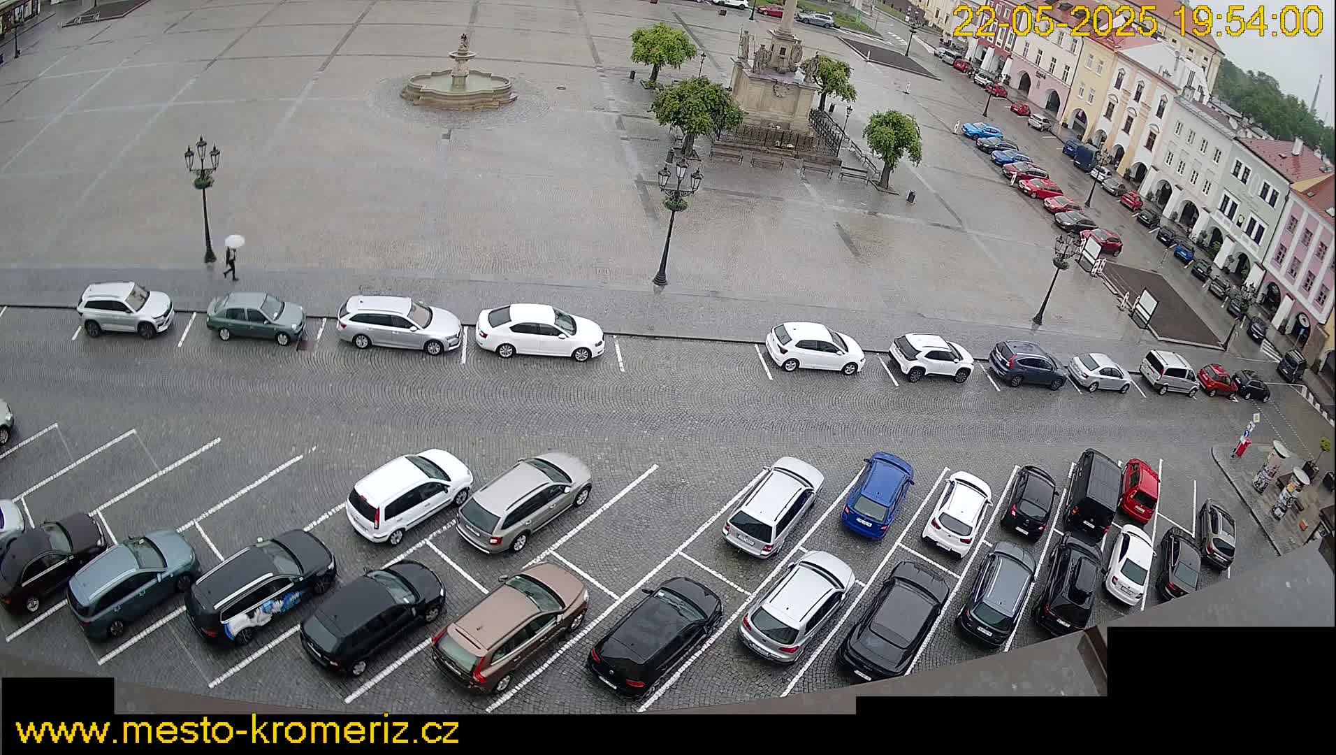 A rain-slicked town square, featuring a fountain and several parked cars, is viewed from above.